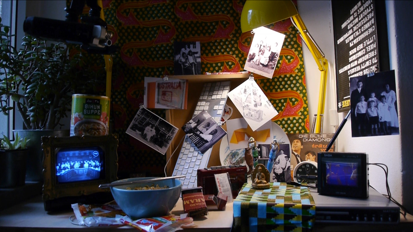 Still from the film "Bagasi", by Meyrick Kaminski. An assortment of objects relating to Indonesia on a table, including textile, media sources, trinkets and cigarette packets.