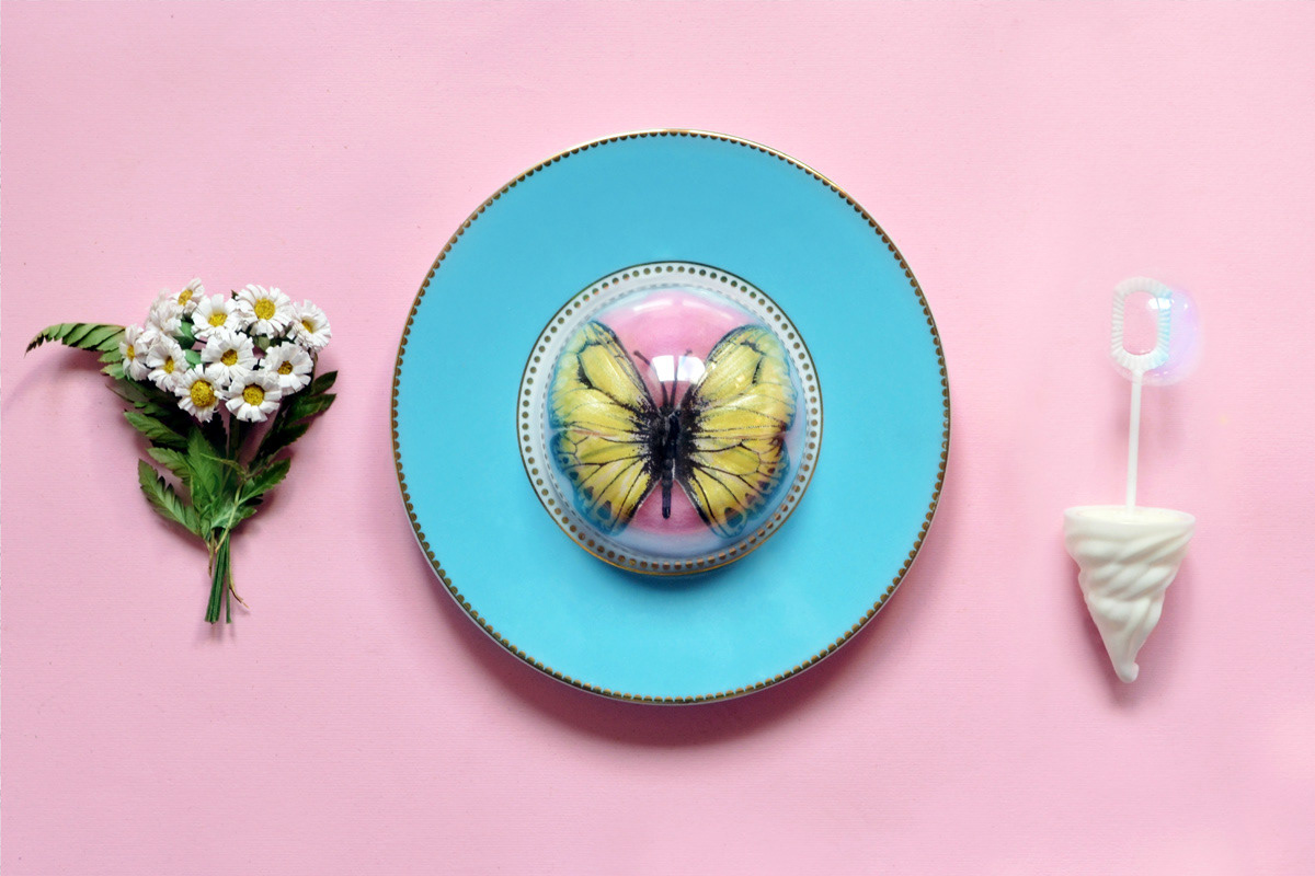 A butterfly encapsulated under a dessert dome on a plate, flanked by artificial flowers and a bubble wand, against a pink background.