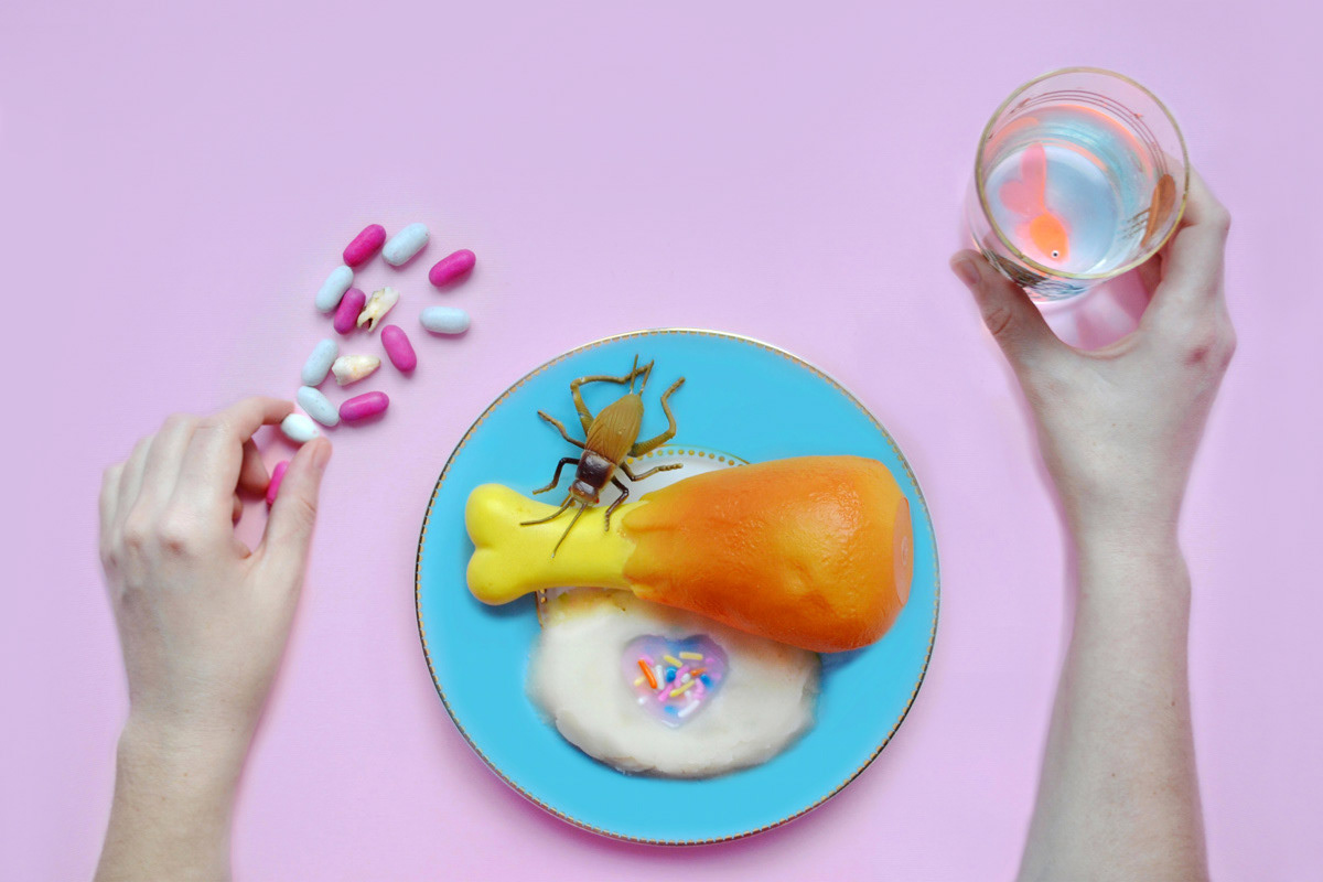 A surreal plate featuring a toy chicken leg, insect, and colorful sprinkles on mashed potato, surrounded by pills, a baby tooth, and a goldfish in a glass of water. Two hands reach in.
