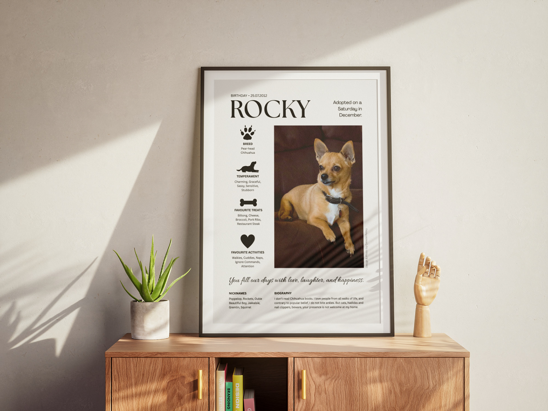 Pet portrait design in a black frame on a table with a plant, wooden hand ornament and books.