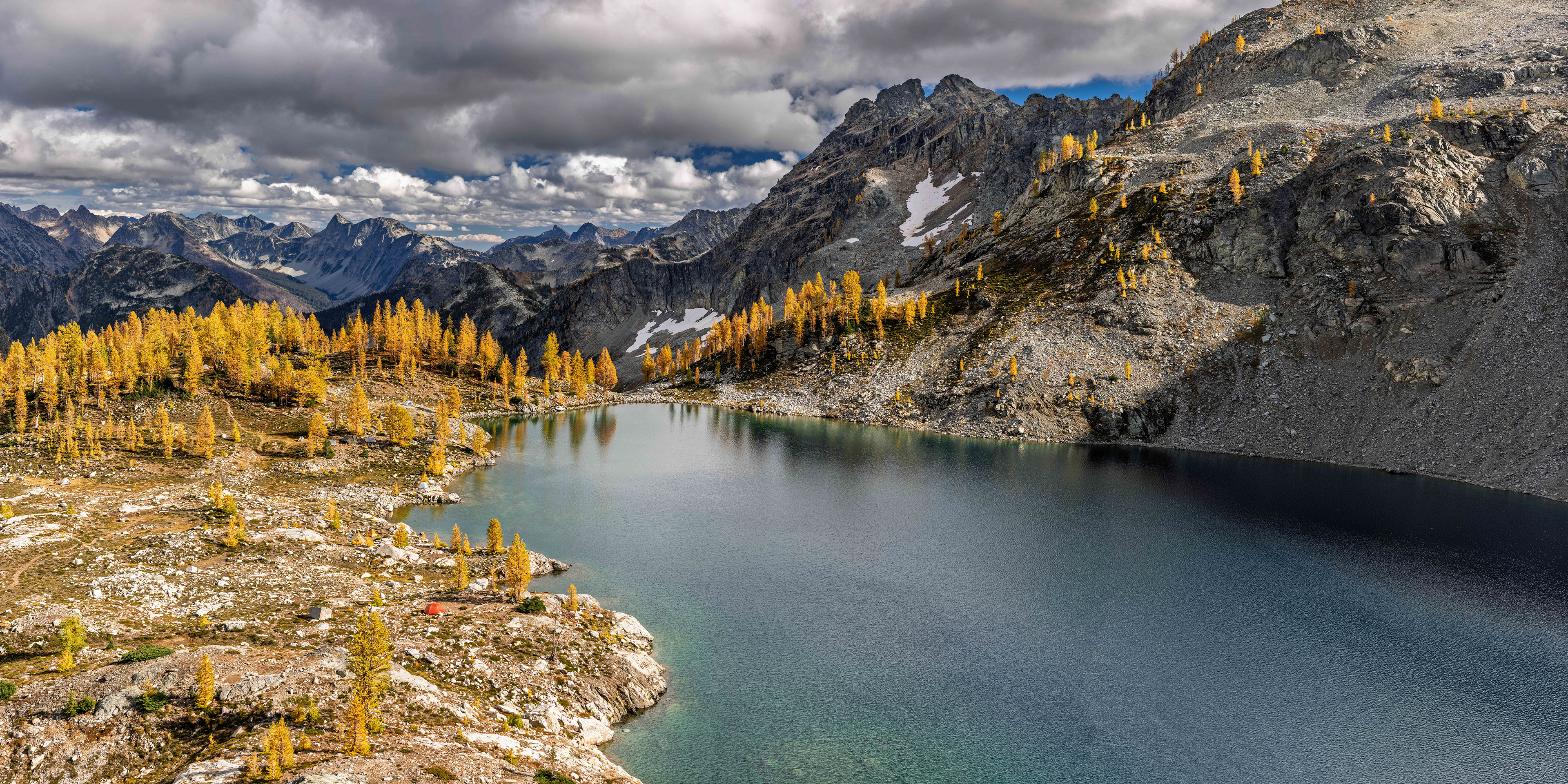 Golden fall larches surround the rocky shoreline of Wing Lake beneath rugged Cascade peaks and dramatic clouds in Washington.