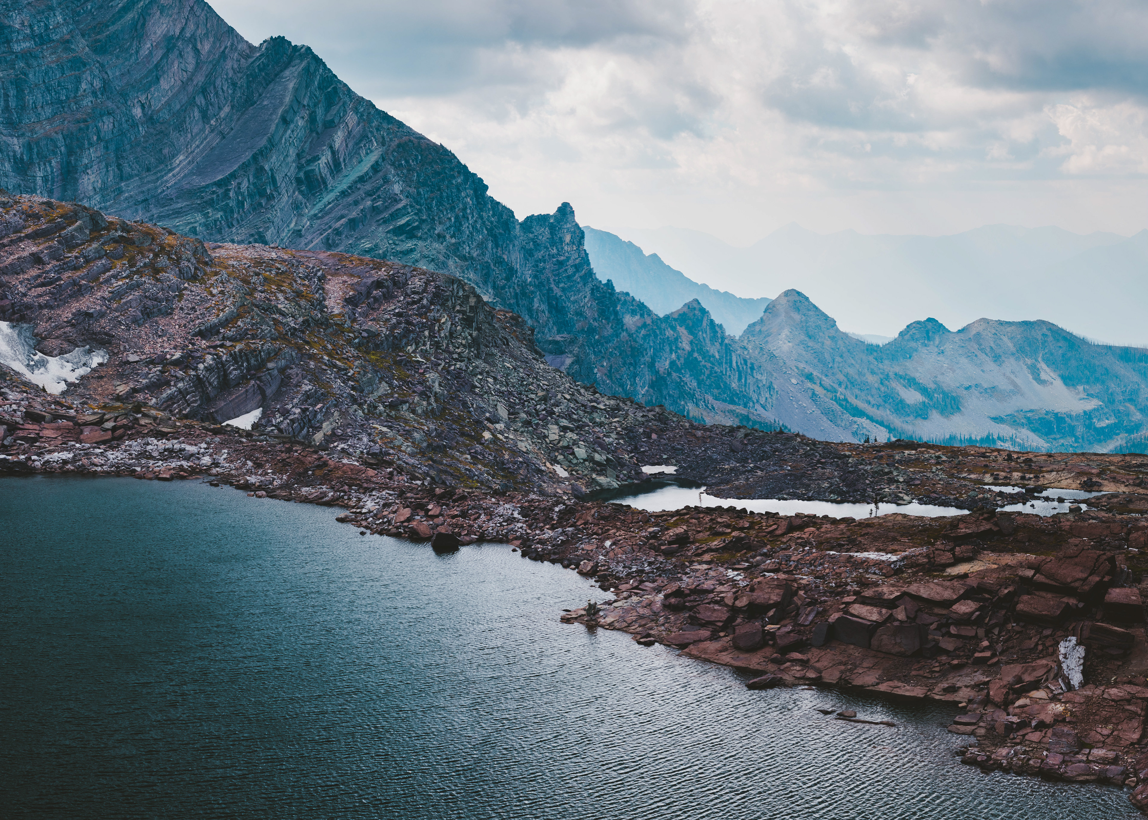 Muted color photograph of Akaiyan Lake in Glacier National Park, featuring a calm alpine lake surrounded by rugged mountain peaks and layered clouds