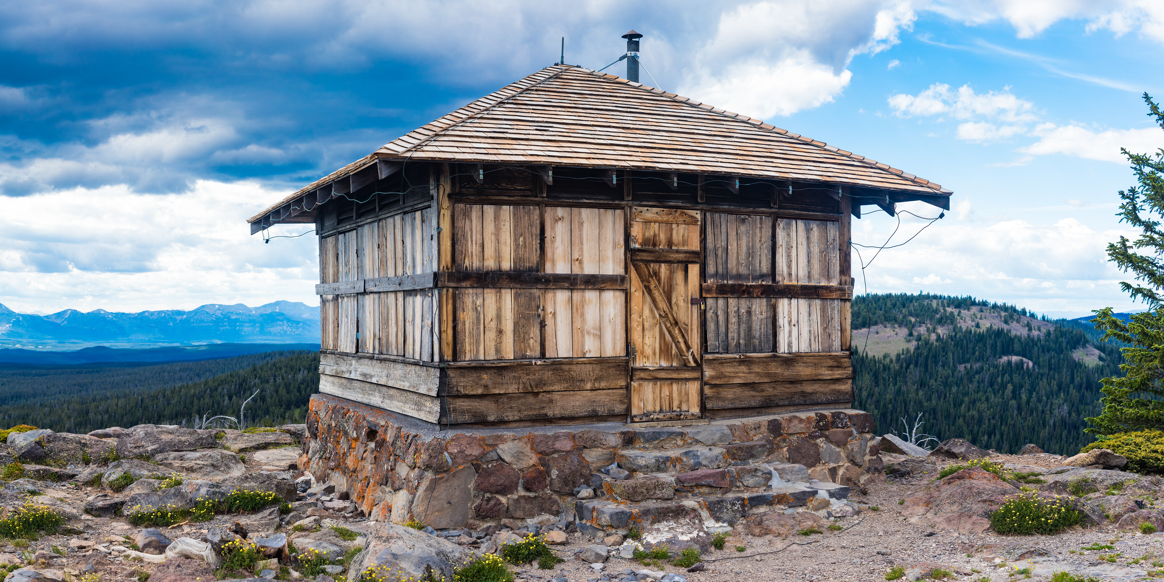 Photograph of the Observation Peak Lookout in Yellowstone National Park, Wyoming, showing a historic wooden fire lookout perched on a rocky summit with sweeping forest and mountain views.