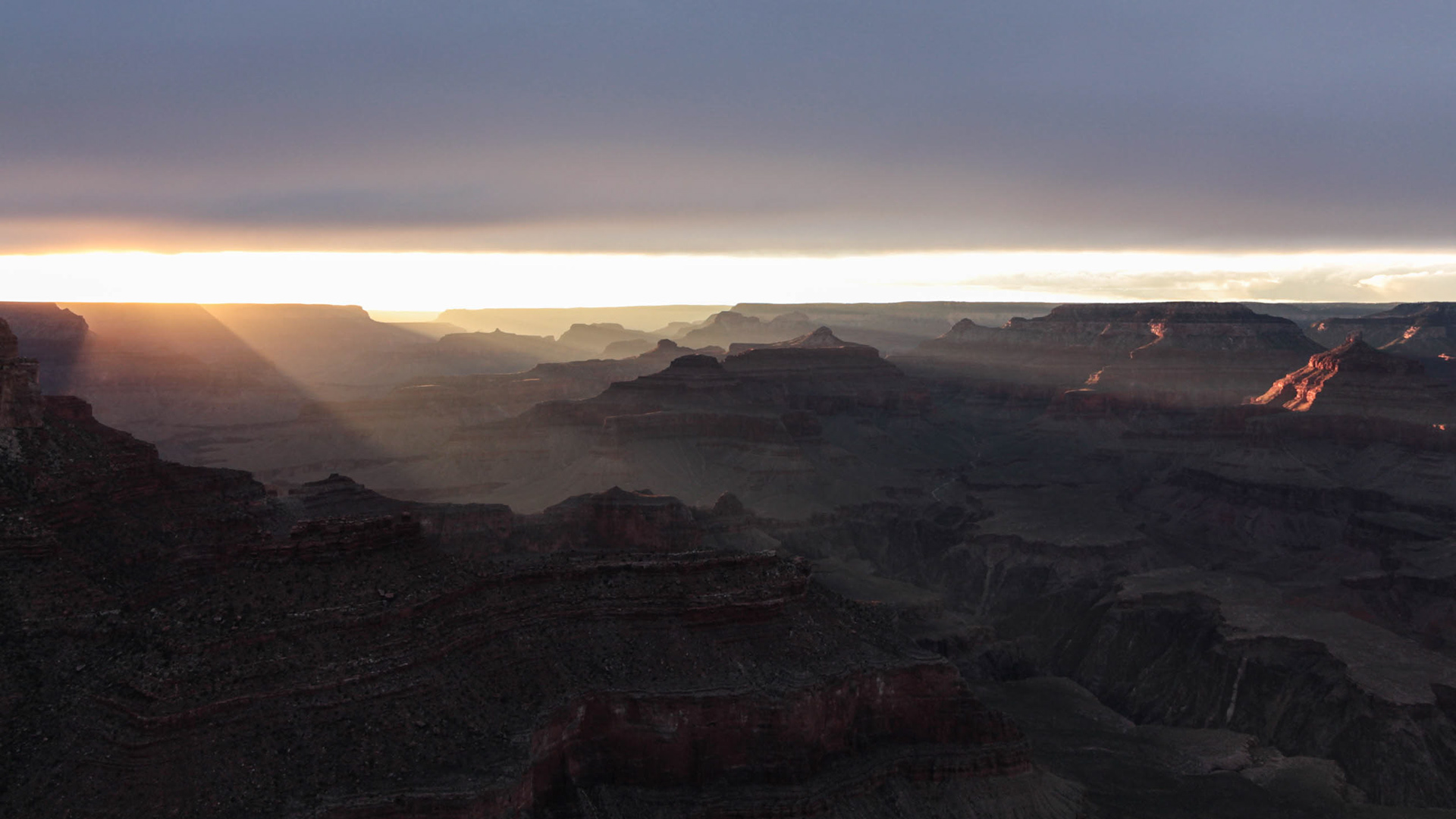 Photograph of sun beams breaking through clouds over the Grand Canyon in Grand Canyon National Park, Arizona, illuminating layered canyon walls and desert terrain.