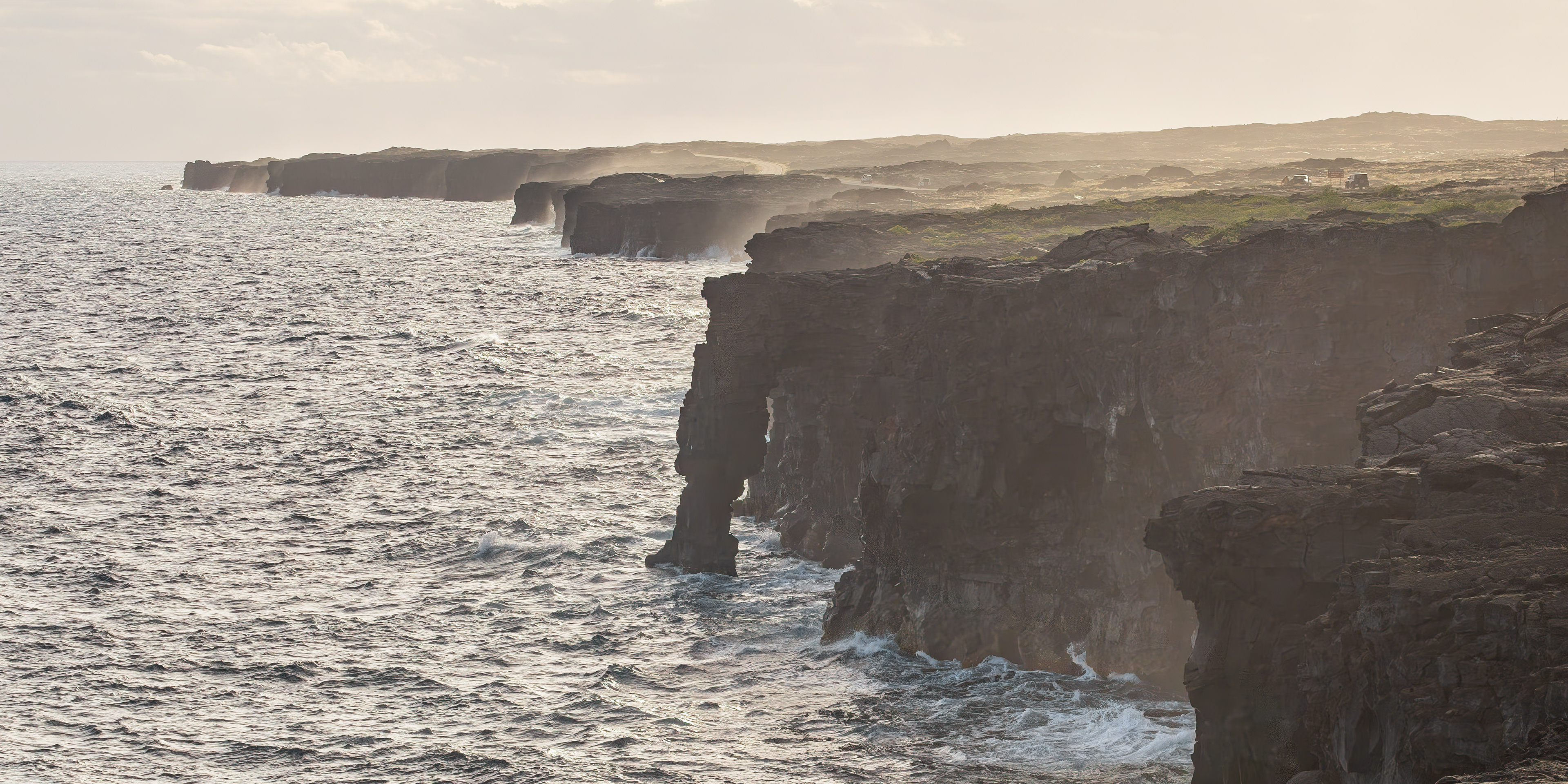 Hōlei Sea Arch illuminated by warm golden-hour light along the rugged volcanic coastline of Hawaiʻi Volcanoes National Park, with the Pacific Ocean crashing against the lava rock below.