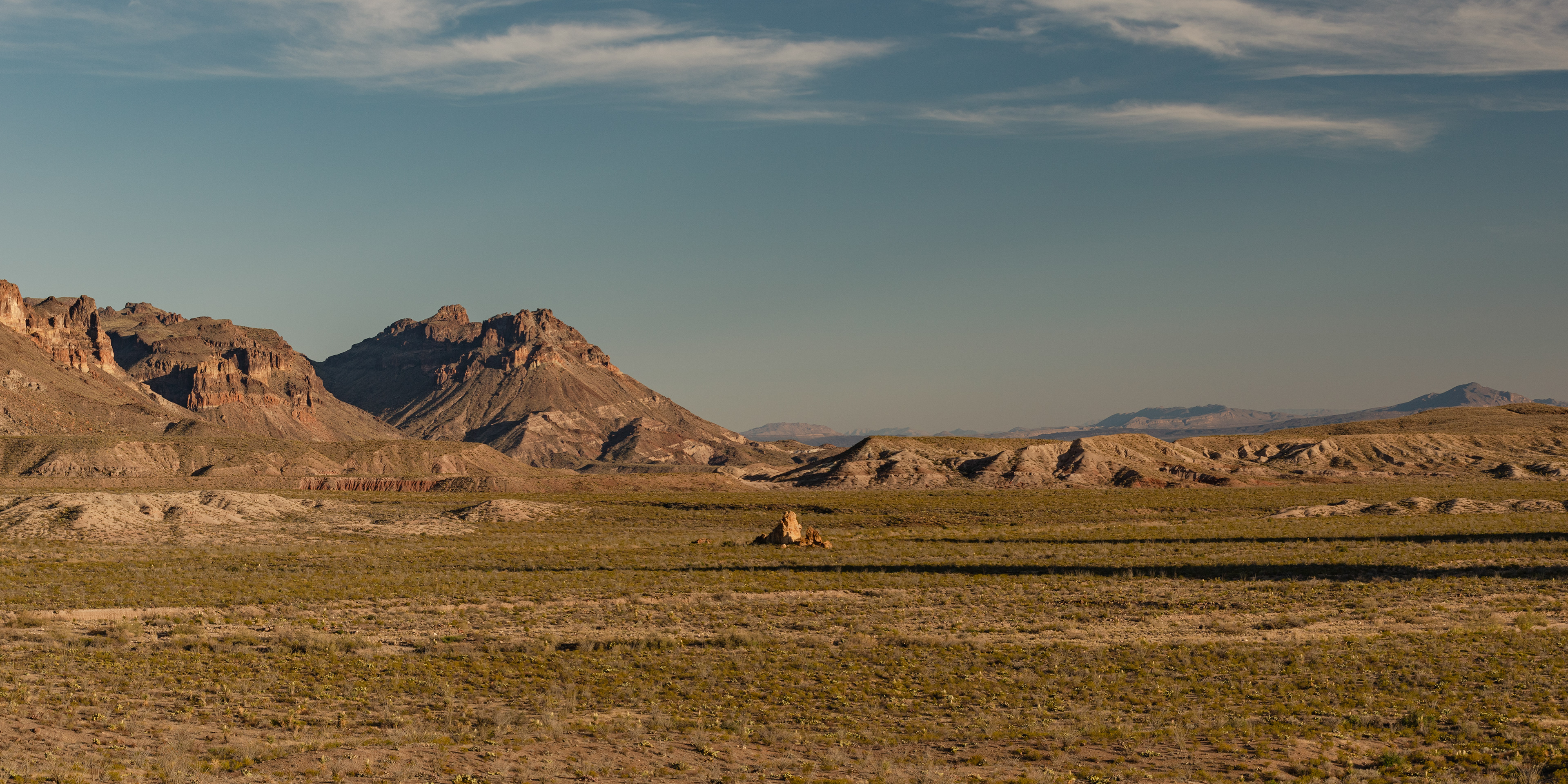 Photograph of the Mule Ears Trail at golden hour in Big Bend National Park, Texas, with warm evening light across desert hills, volcanic rock formations, and arid vegetation.
