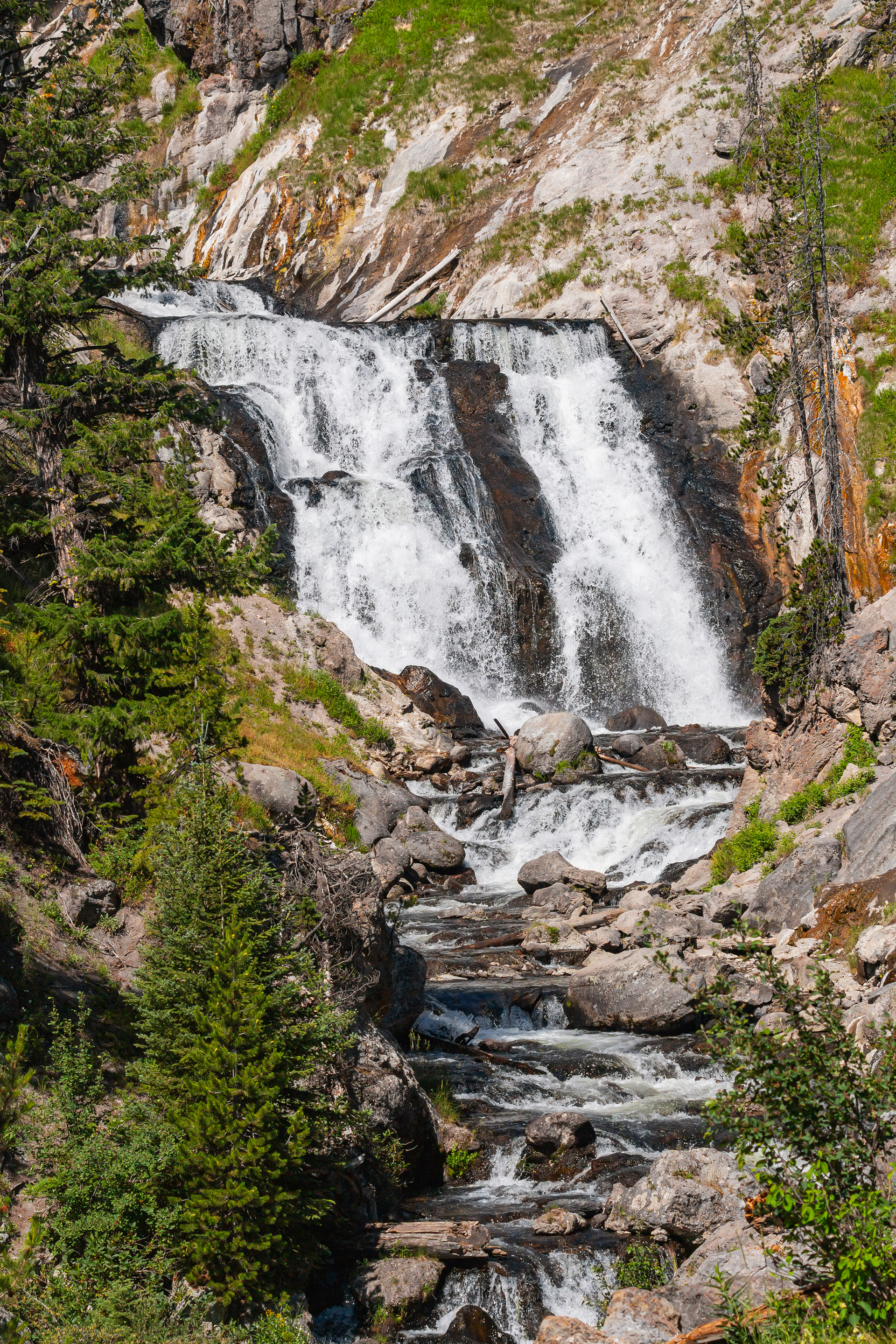 Photograph of Wraith Falls in Yellowstone National Park, Wyoming, with a tall waterfall cascading down a rugged cliff face surrounded by forested slopes.