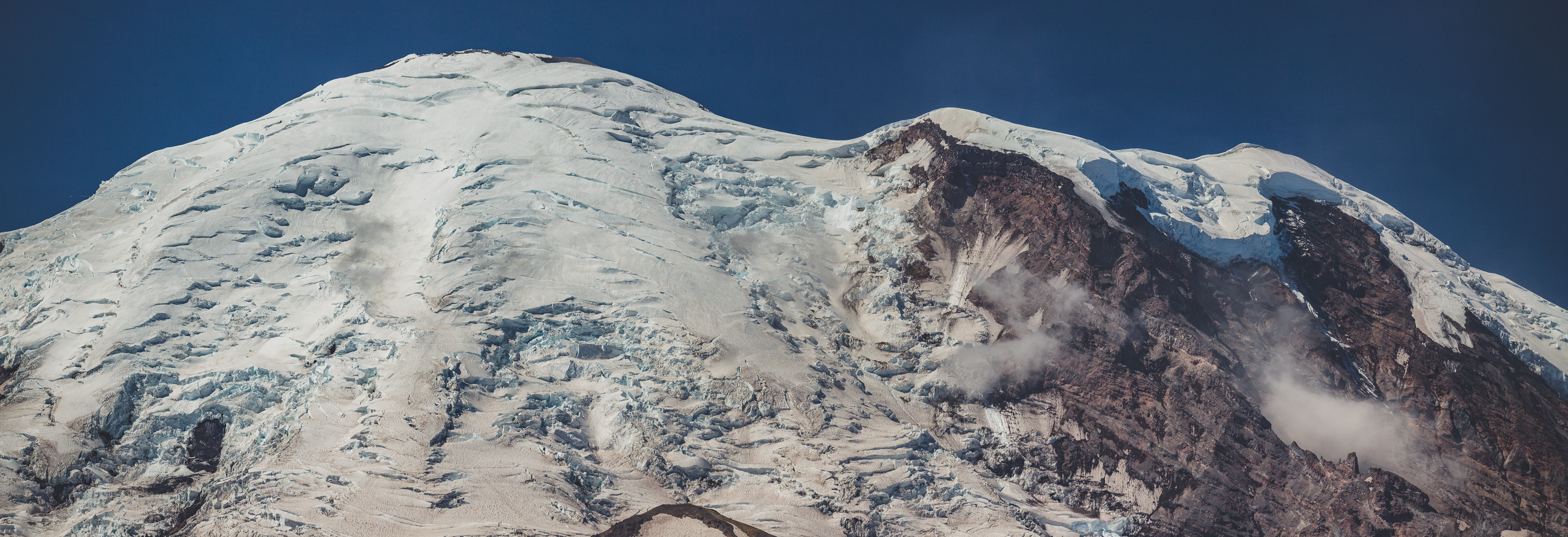 View of Mount Rainier from the Burroughs Mountain Trail, with snow-covered volcanic slopes rising above alpine terrain and rocky ridgelines under a clear sky