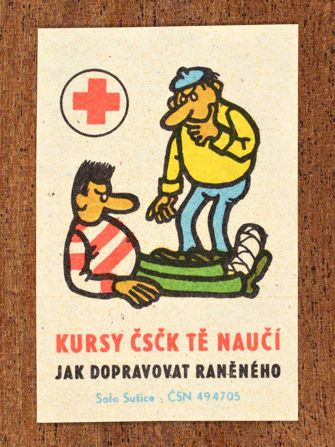 Vintage c. 1966 Czech matchbox label showing a man assisting another injured man lying on the floor with a bandaged foot. Produced by the Solo Sušice match factory to promote first-aid education and correct methods of transporting injured persons in cooperation with the Czechoslovak Red Cross.