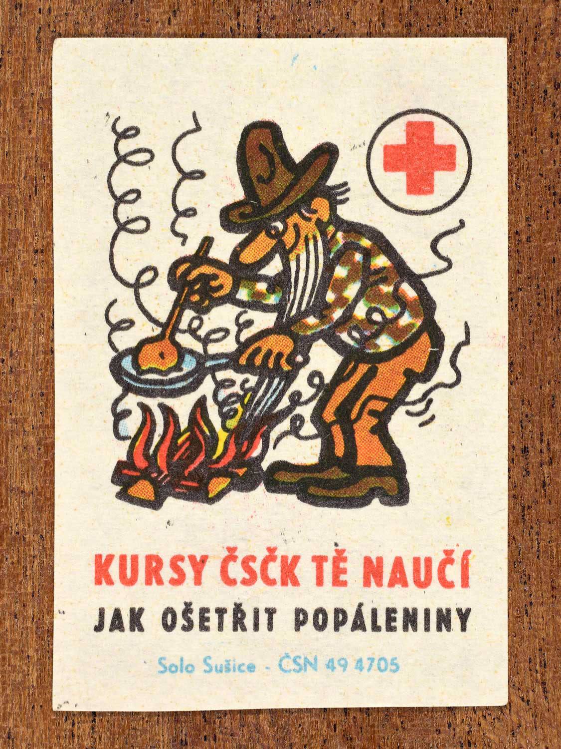 Vintage c. 1966 Czech matchbox label showing a man cooking food over a campfire fire. Produced by the Solo Sušice match factory to promote first-aid education and proper treatment of burns in cooperation with the Czechoslovak Red Cross.