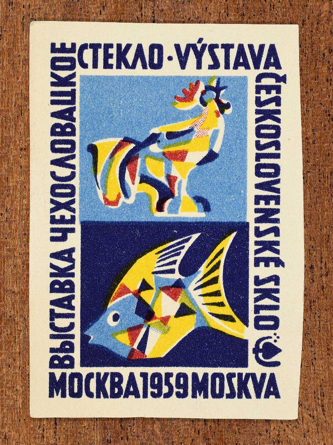 An abstract phillumeny graphic featuring a stylised bird and ﬁsh in blue, yellow, and red depicted on a 1959 Czechoslovak matchbox label by an unknown factory. The design functions to showcase animal figurines created from coloured glass and exhibited at an event in Moscow.