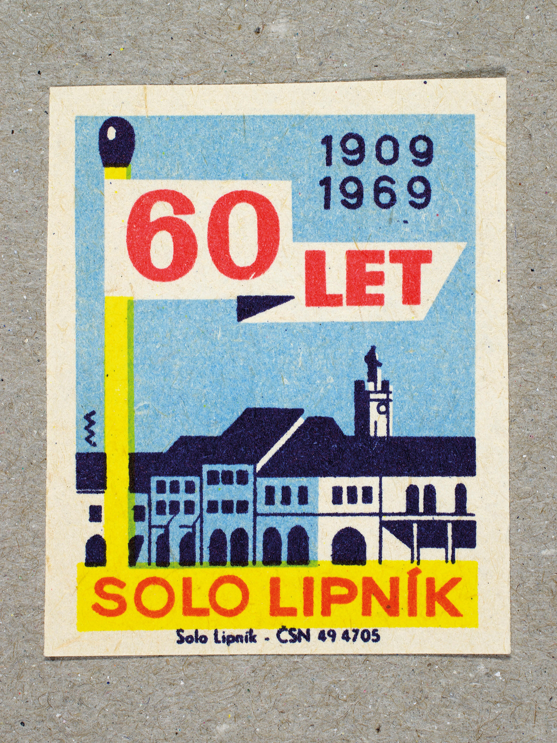 Vintage 1969 Czech matchbox label by Solo Lipník featuring a match as a flag pole standing over a factory, in blue and yellow colours. Part of the 'SOLO Lipník Anniversary' series advertising the Czechoslovakian match factory.