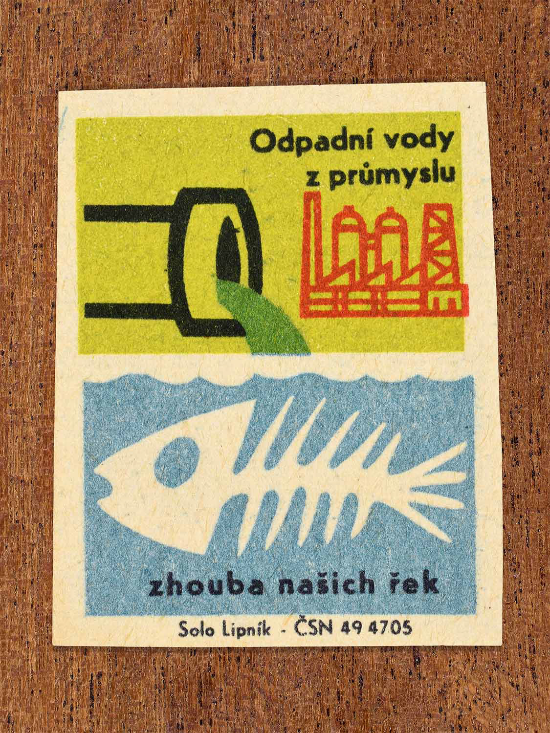 Vintage c. 1966 Czech matchbox label showing a factory pipe discharging green industrial waste into water above a fish skeleton in green, blue and red colours. Produced by the Solo Lipník match factory to highlight that industrial wastewater destroys rivers.