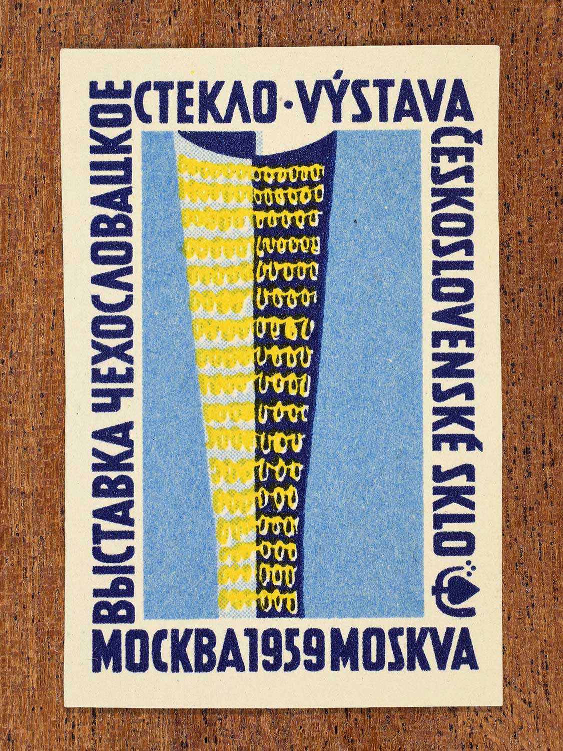 Phillumeny graphic featuring a blue and yellow glass vessel on a light blue background. Depicted on a 1959 Czechoslovak matchbox label, the design functions to showcase Czechoslovak glass craftsmanship as exhibited at an event in Moscow.