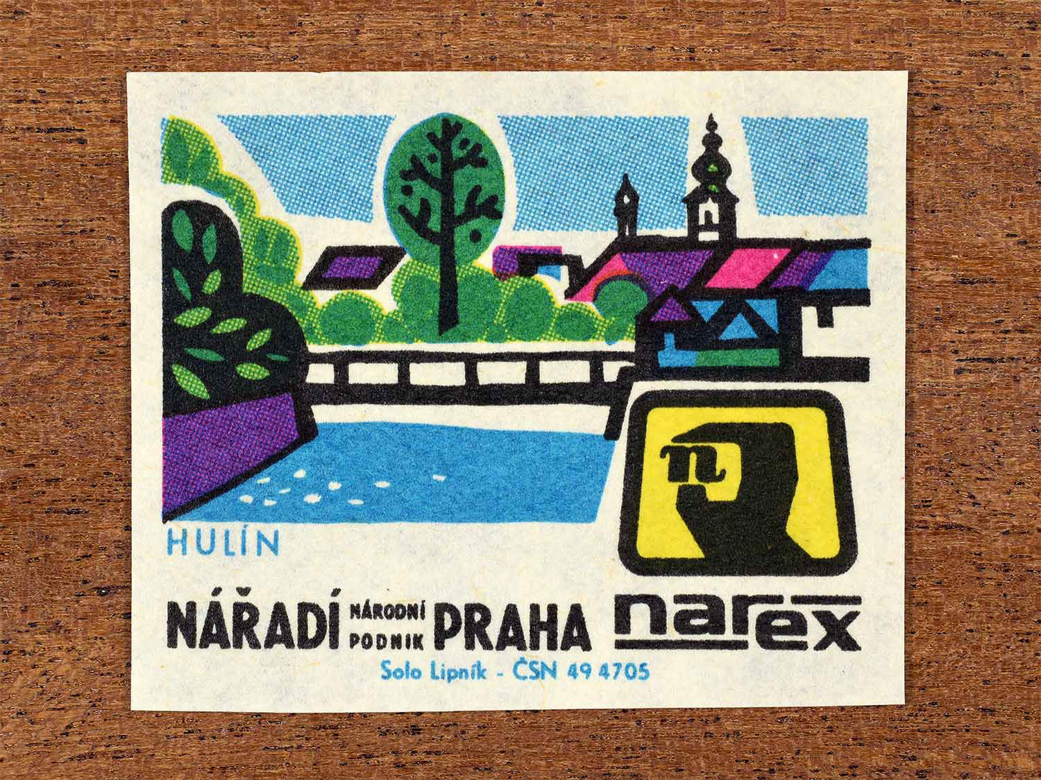 Vintage 1967 Czech matchbox label showing a riverside town with bridge, trees, and church towers in multiple bright coours. Produced by the Solo Lipník match factory to promote Hulín and Narex, the national tool enterprise in Czechoslovakia.