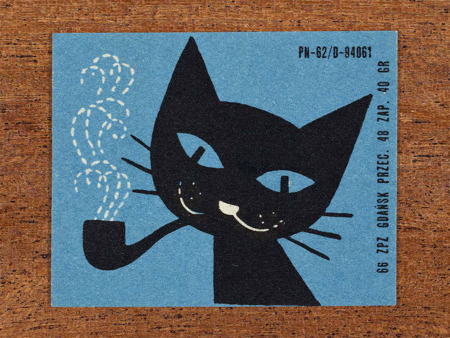 Vintage 1966 Polish matchbox label showing a stylised white cat smoking a pipe with white dotted smoke on a blue background. Produced by the Gdańsk match factory in Poland as retail packaging for safety matches.