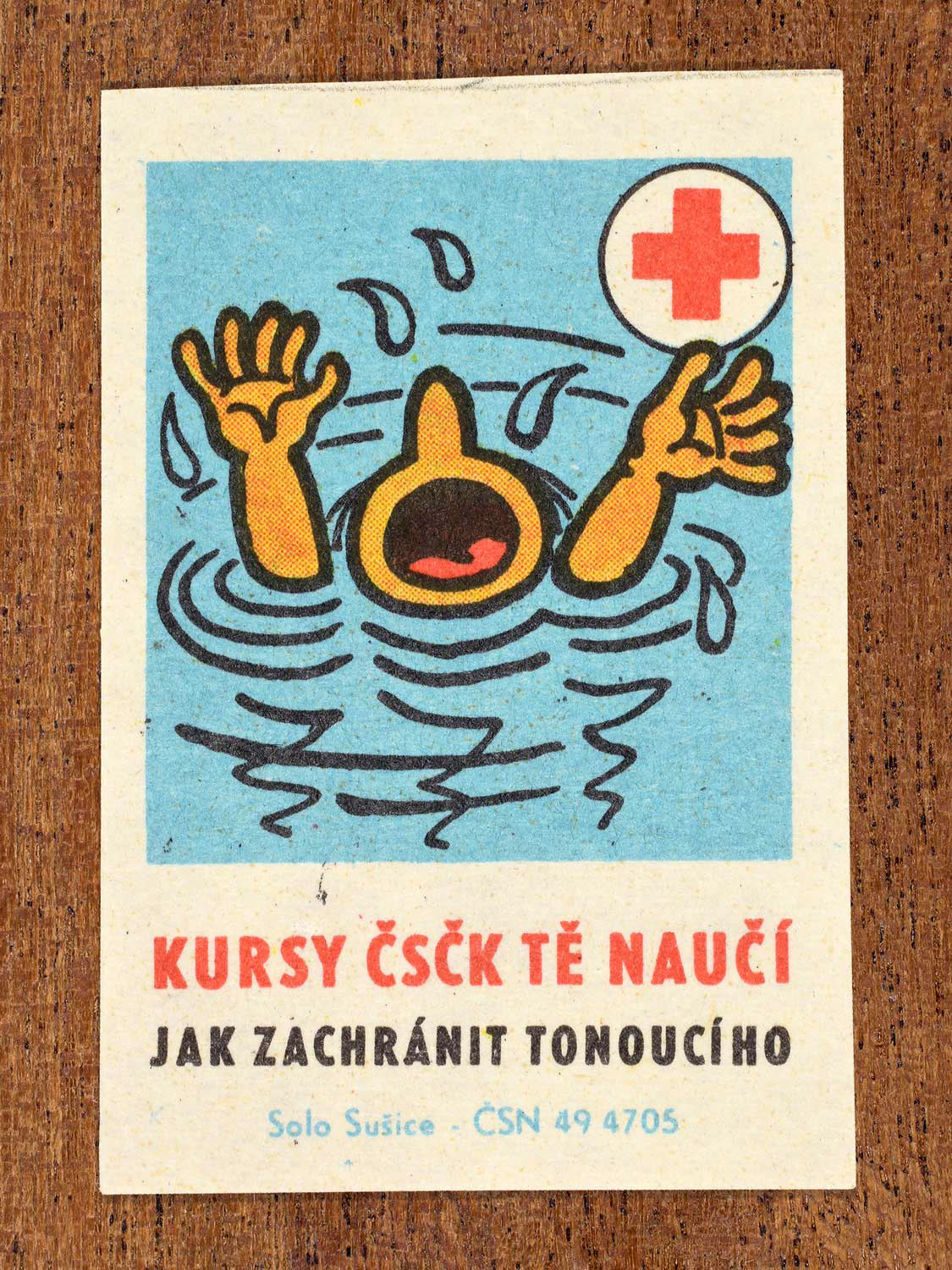 Vintage c. 1966 Czech matchbox label showing a drowning person with raised arms struggling in water. Produced by the Solo Sušice match factory to promote first-aid education and water rescue awareness in cooperation with the Czechoslovak Red Cross.