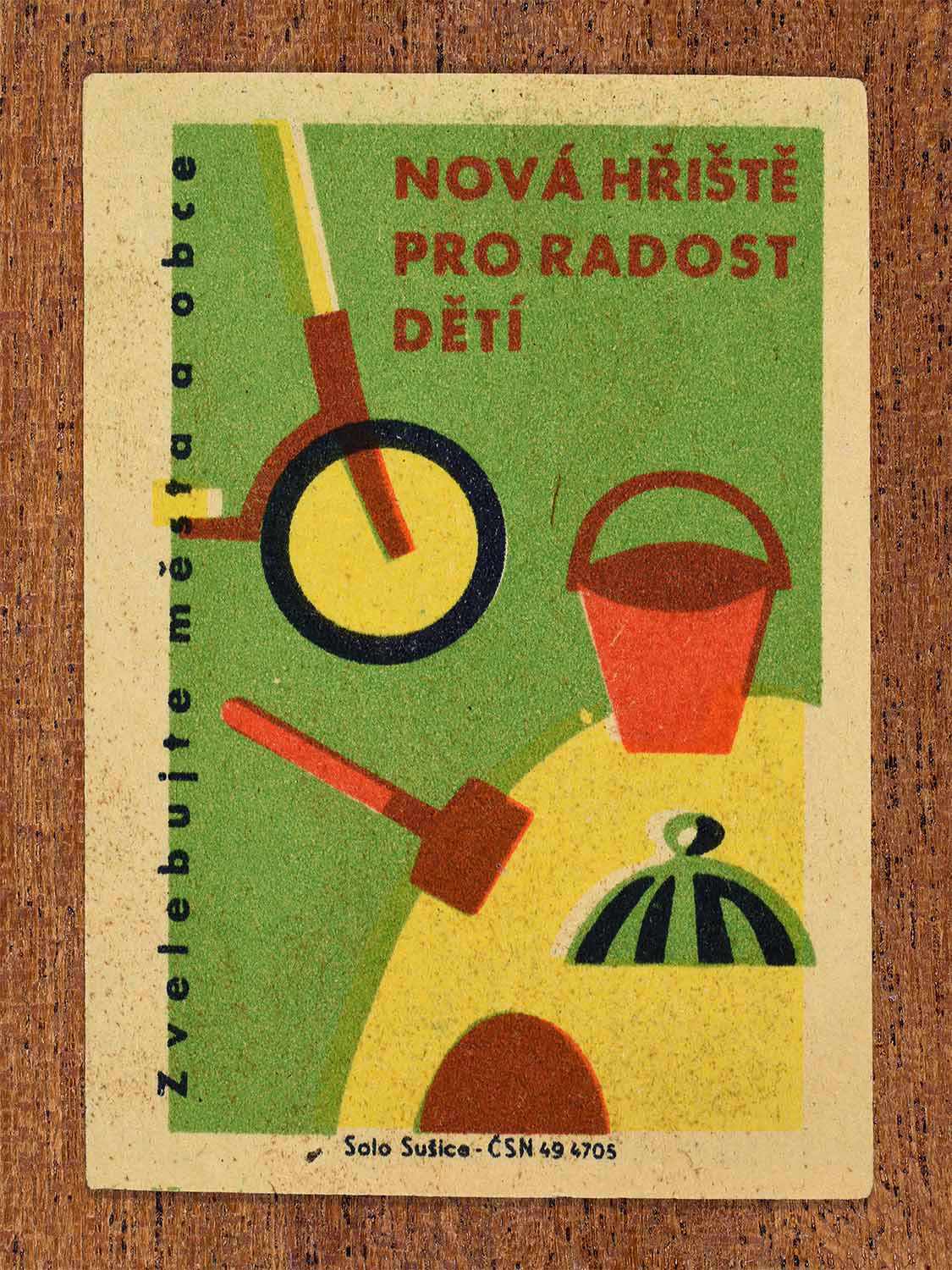 Vintage 1961 Czech matchbox label showing a playground setting with a scooter, sand pit and bucket, on a green background. Produced by the Solo Sušice match factory to encourage building new playgrounds for the benefit of children.