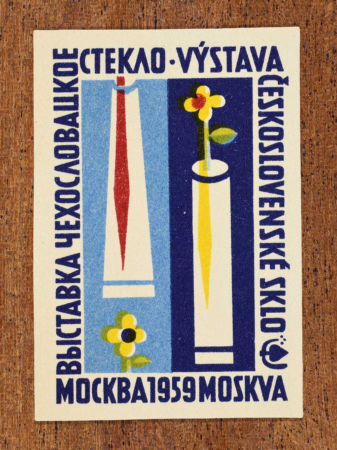 Mid-century modern Eastern Bloc illustration on a 1959 Czechoslovak matchbox label featuring two glass vessels, one holding a single yellow ﬂower on a dark blue background. Designed to promote an international glass exhibition in Moscow by an unknown factory.