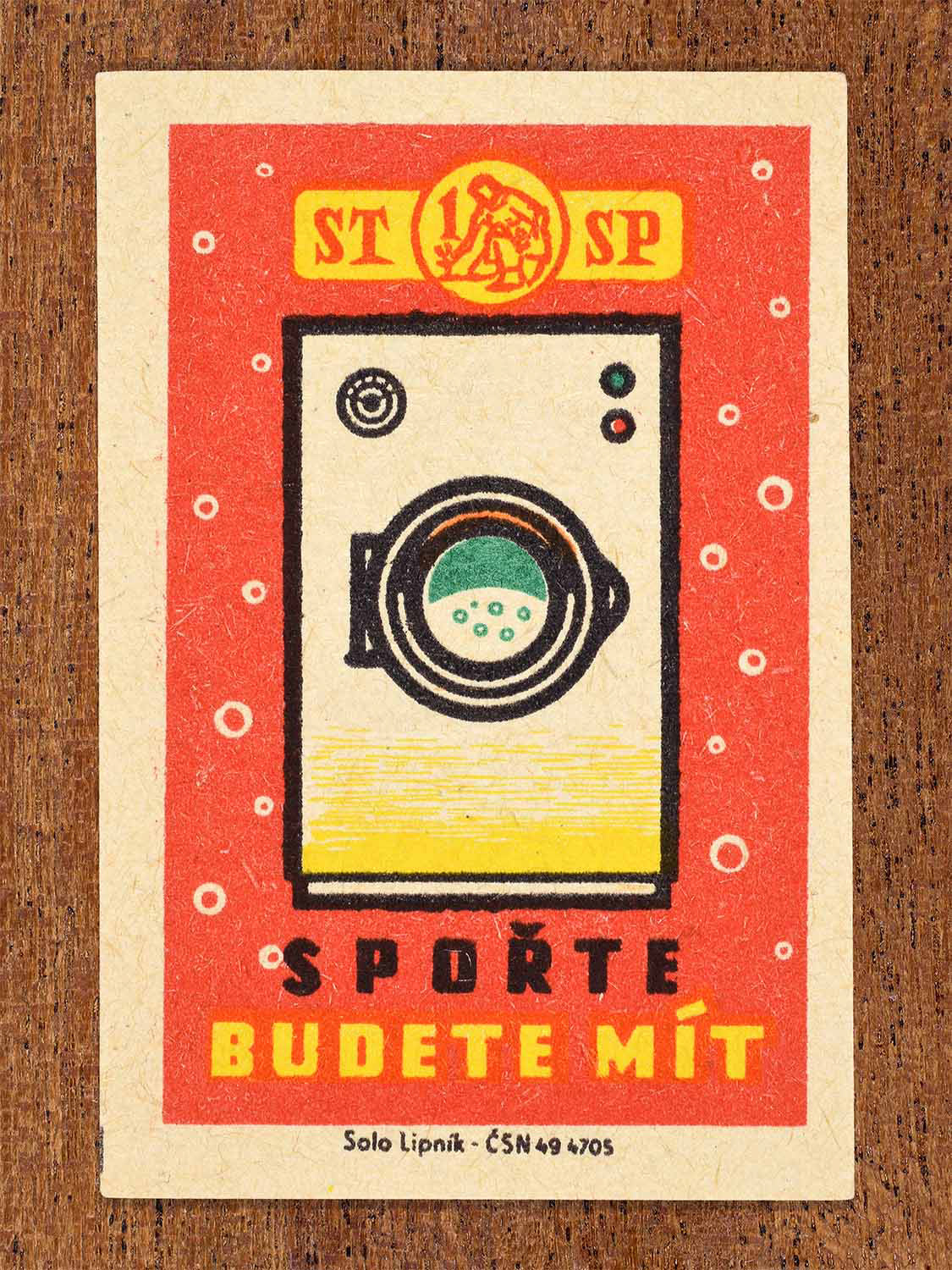 Vintage 1960 Czech matchbox label showing a front-loading washing machine on red background. Produced by the Solo Lipník match factory to promote saving in order to afford modern household goods.
