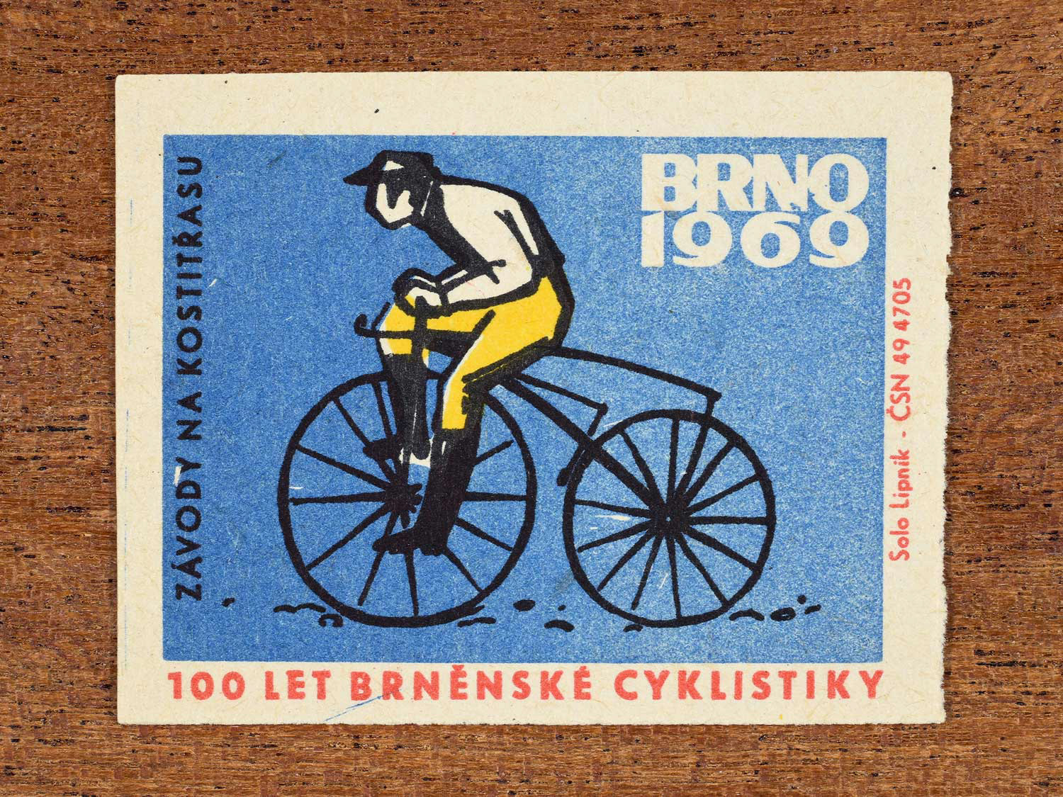Vintage 1969 Czech matchbox label showing a stylised cyclist on an antique bicycle, on a blue background. Produced by the Solo Lipník match factory to celebrate 100 years of cycling in Brno, Czechoslovakia.