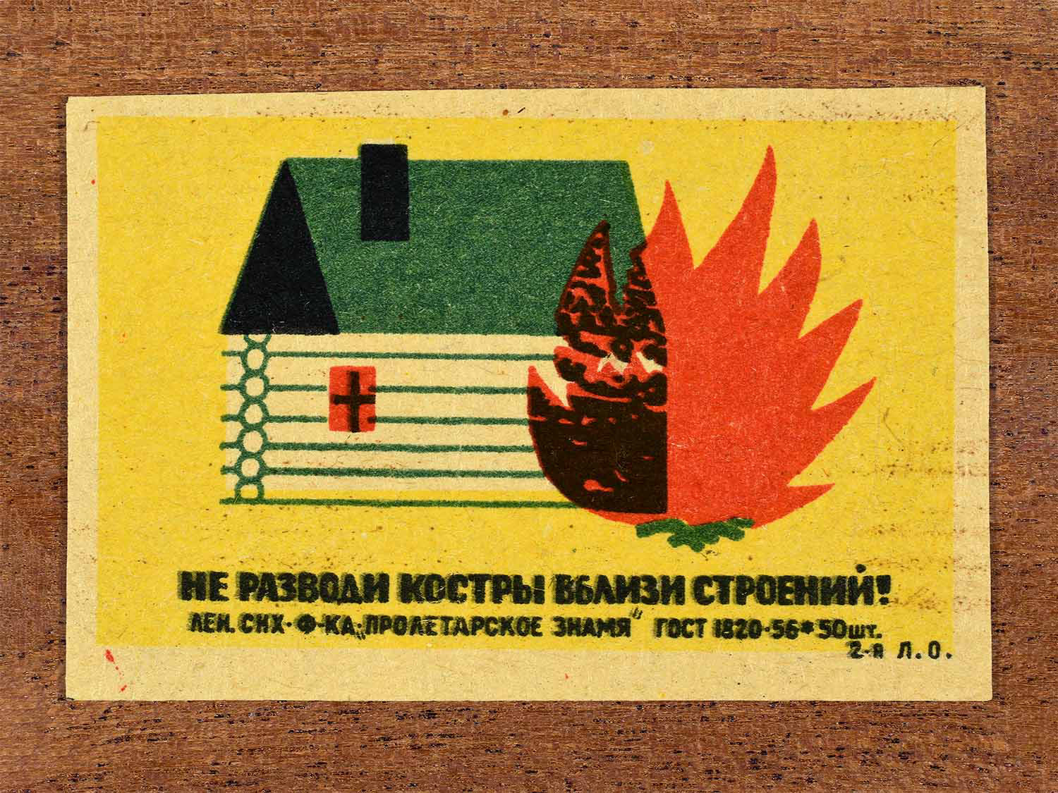 Vintage 1962 Soviet matchbox label showing a campfire next to a wooden house warning against open flames. In yellow, green, yellow and black colours. Produced by the Proletarian Banner match factory to promote safe distance from buildings.