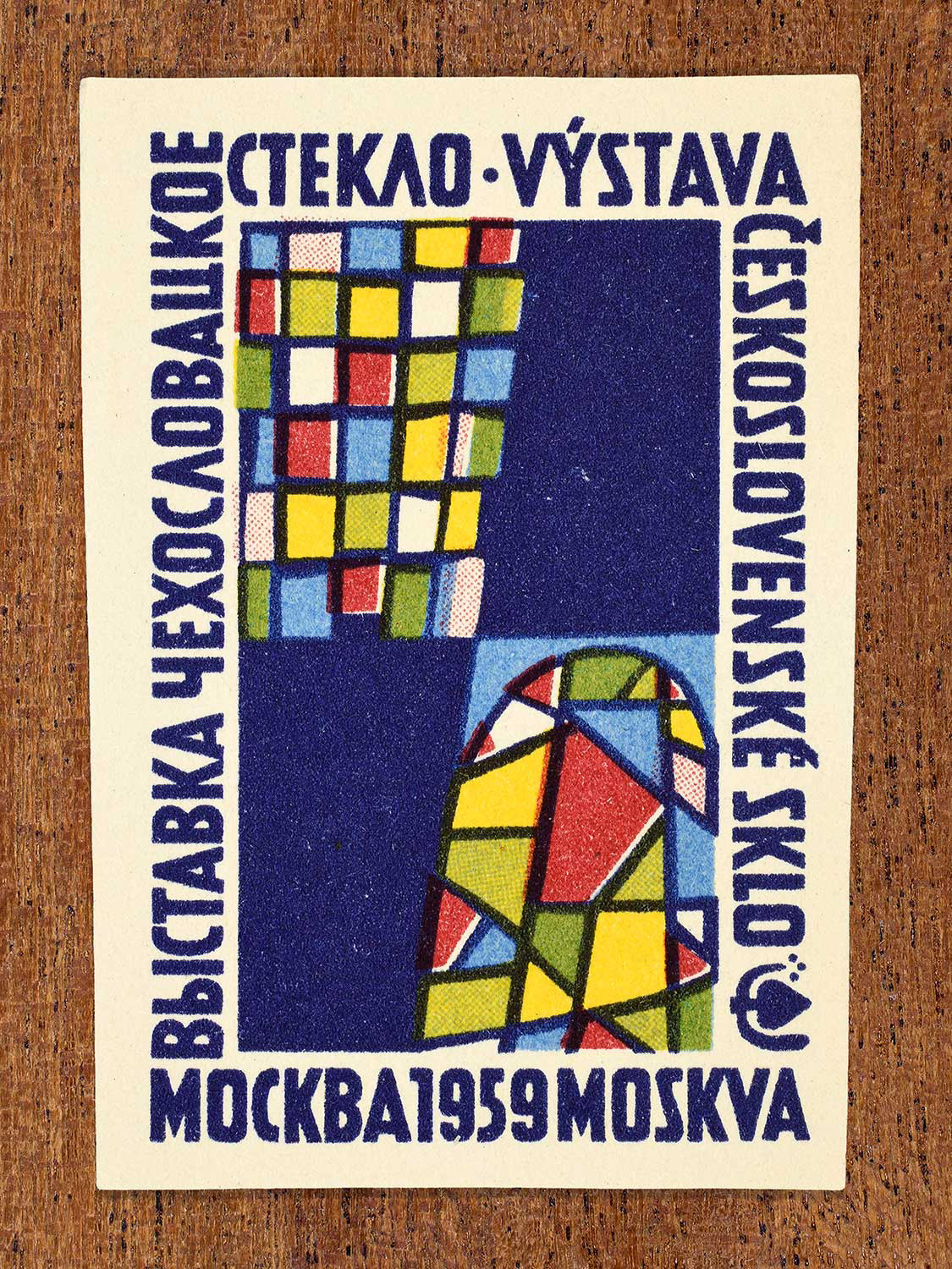 An illustration of a multi-coloured geometric glass sculpture on a blue background depicted on a vintage Czech matchbox label. The design functions to promote an international glass exhibition in Moscow, 1959.