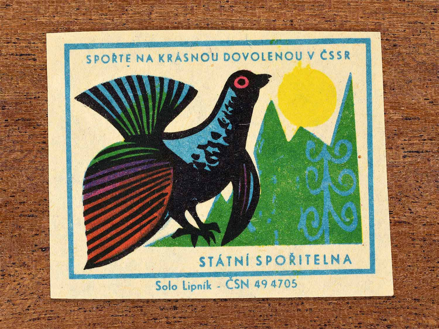 Vintage 1968 Czech matchbox label showing a stylised bird displaying in a forest landscape beneath the sun. Produced by the Solo Lipník match factory to promote saving money for holidays in the Czechoslovak Socialist Republic.
