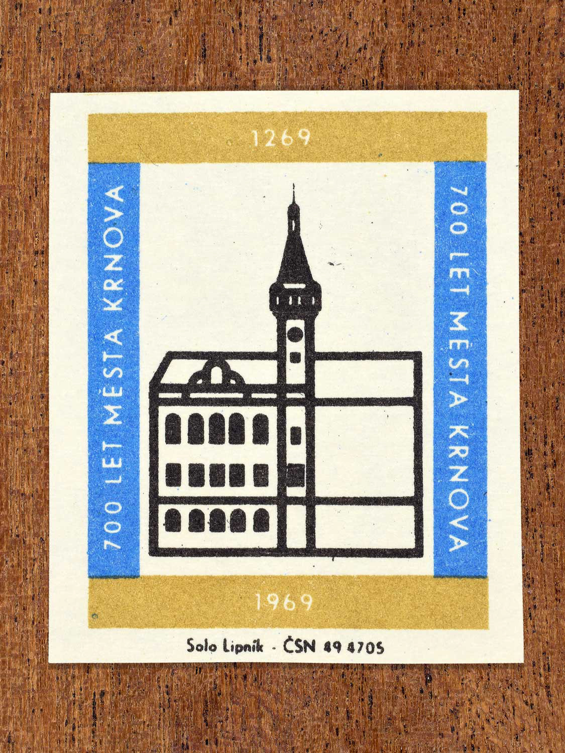 Vintage 1969 Czech matchbox label showing a historic civic building of Krnov with a prominent tower, in black with blue and gold framing. Produced by the Solo Lipník match factory to commemorate 700 years of the city of Krnov.