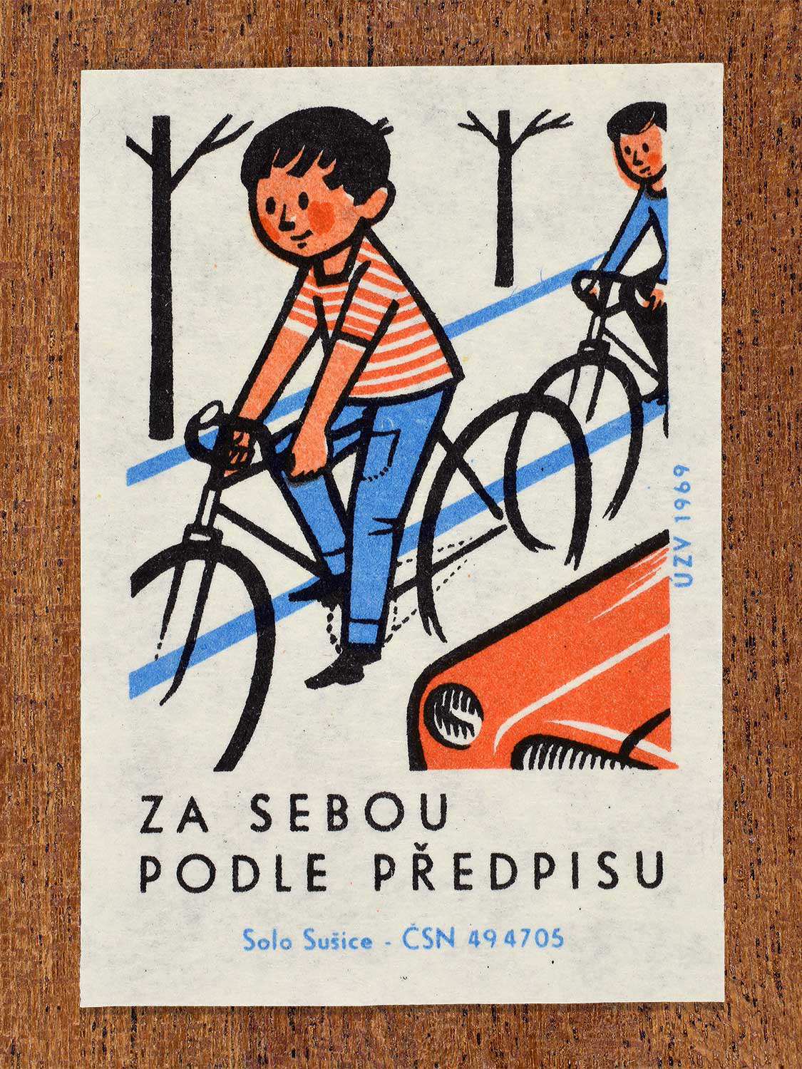 Vintage 1969 Czech matchbox label showing two children riding bicycles in a single file line on the side of a road near a red car. Produced by the Solo Sušice match factory to promote safe cycling regulations among children.