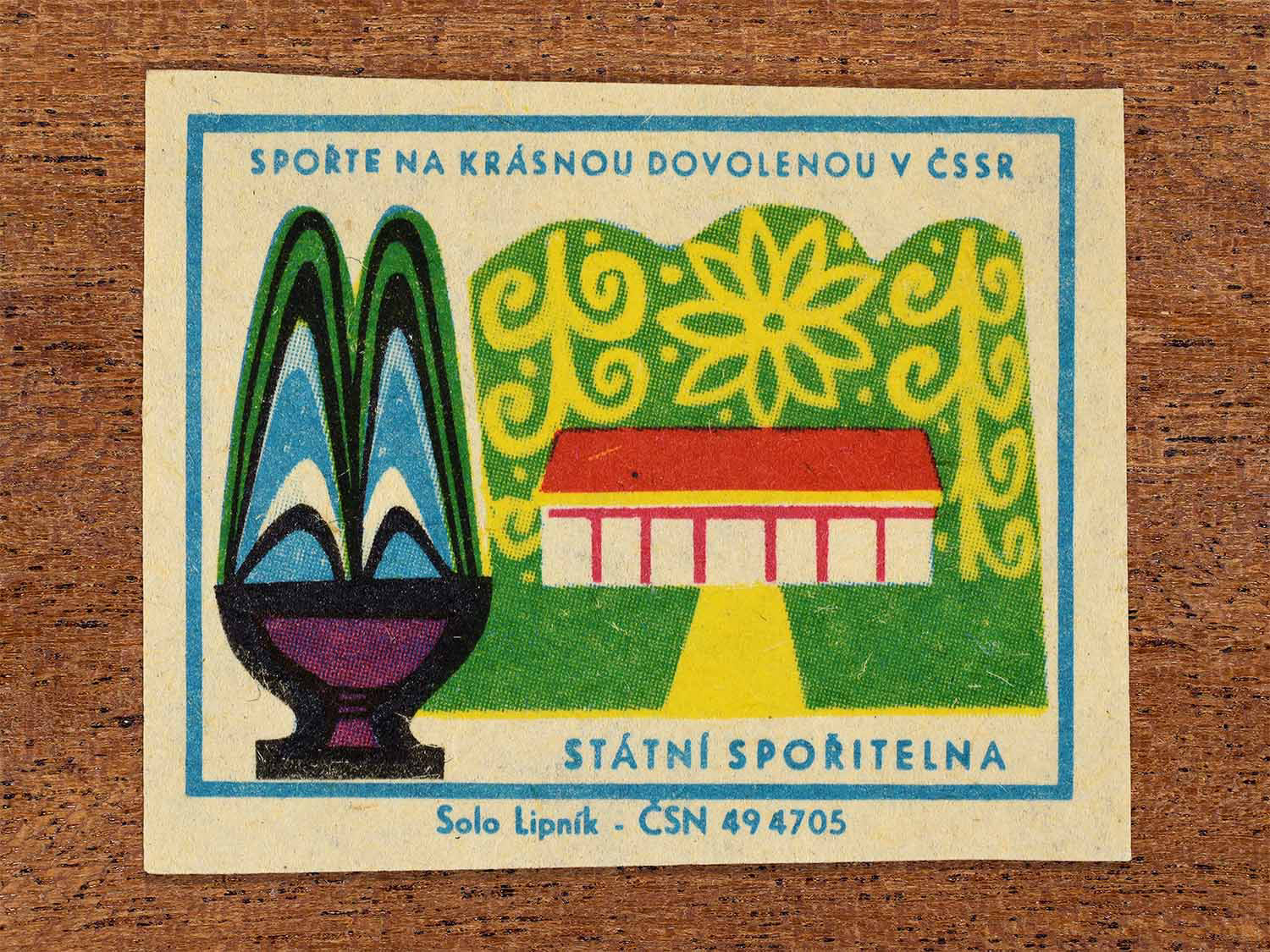 Vintage 1968 Czech matchbox label showing a stylised water fountain next to a building surrounded by trees. Produced by the Solo Lipník match factory to promote saving money for holidays in the Czechoslovak Socialist Republic.