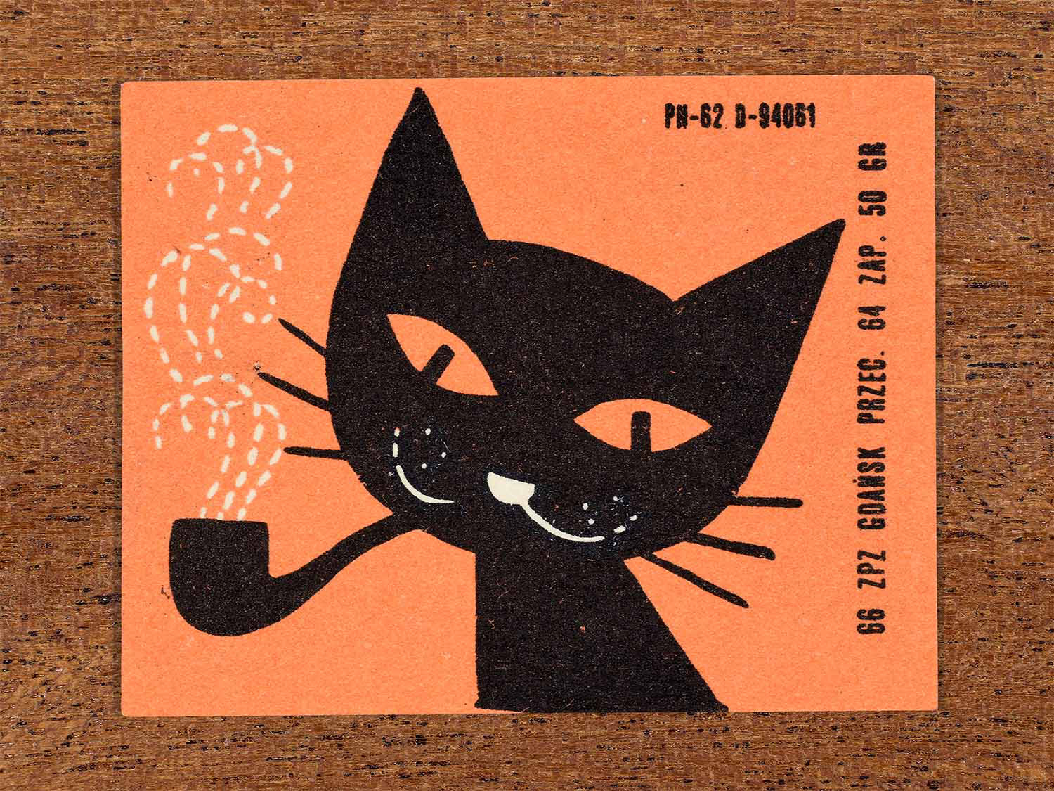 Vintage 1966 Polish matchbox label showing a stylised black cat smoking a pipe with white dotted smoke on a peach background. Produced by the Gdańsk match factory in Poland as retail packaging for safety matches.