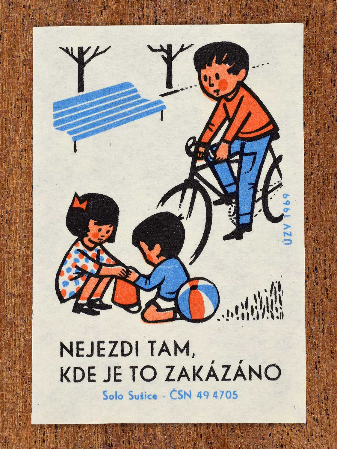 Vintage 1969 Czech matchbox label showing a boy riding a bicycle past a park bench where a girl and boy sits playing. Produced by the Solo Sušice match factory to promote safe and rule-abiding bicycle riding.