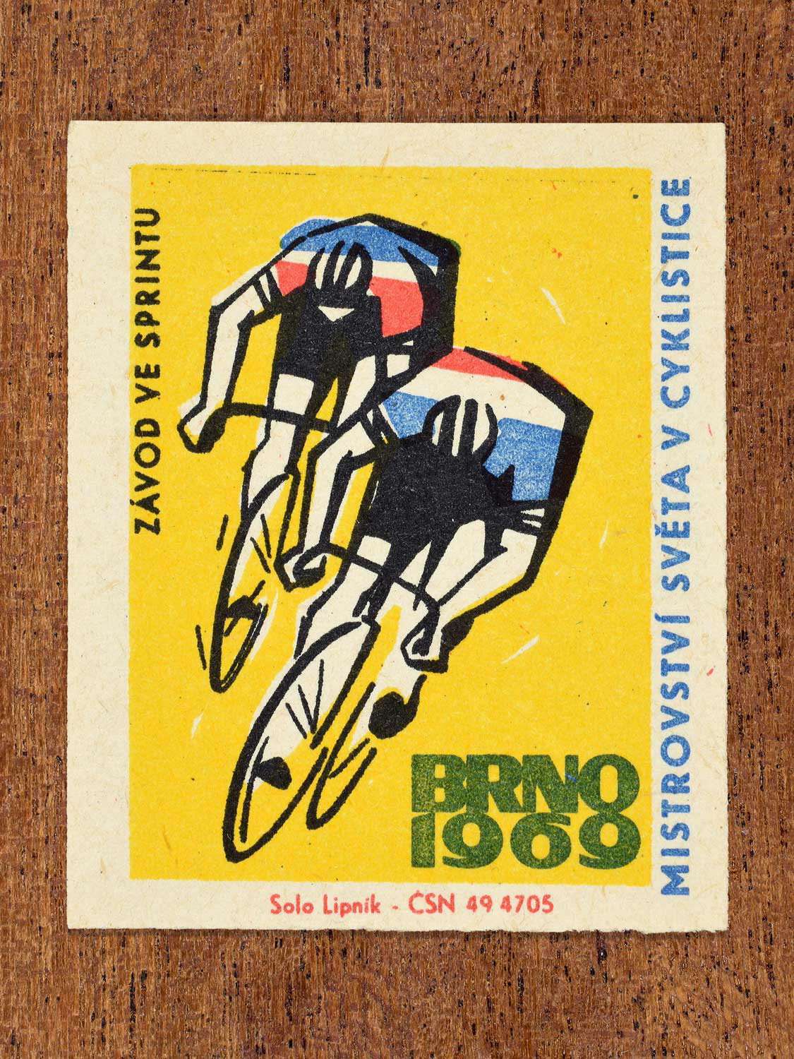 Vintage 1969 Czech matchbox label showing two stylised cyclists competing in a sprint race, on a yellow background. Produced by the Solo Lipník match factory to promote sprint events at a cycling championship in Brno, Czechoslovakia.