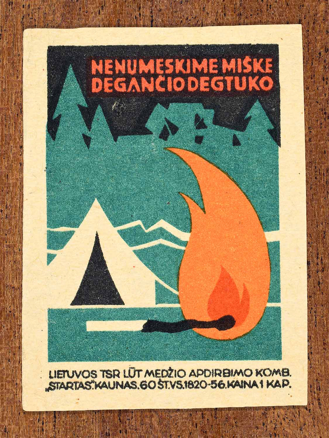 Vintage 1964 Lithuanian matchbox label showing a lit match and flame near forest trees and a tent. In black, green and orange colours. Produced by the Startas match factory to promote fire safety in forests.