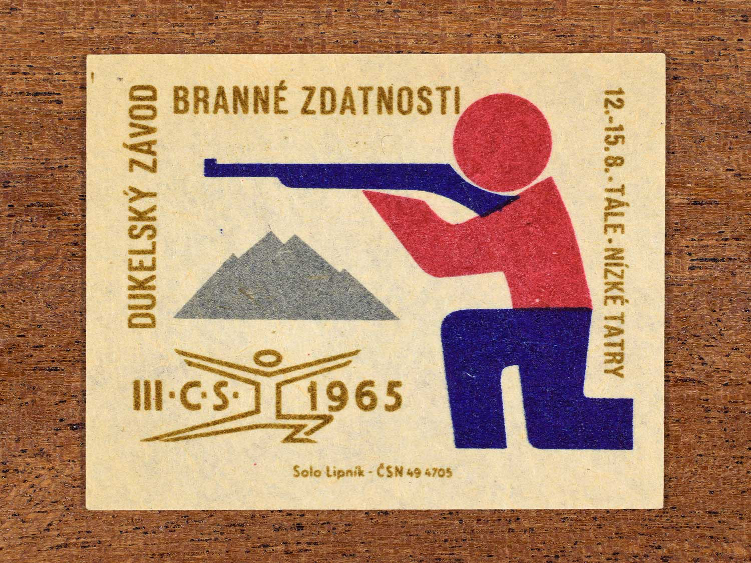 Vintage 1965 Czechoslovak matchbox label showing an abstract figure firing a rifle. In blue, red and gold colours. Produced by the Solo Lipník match factory to promote physical fitness and sporting achievement.