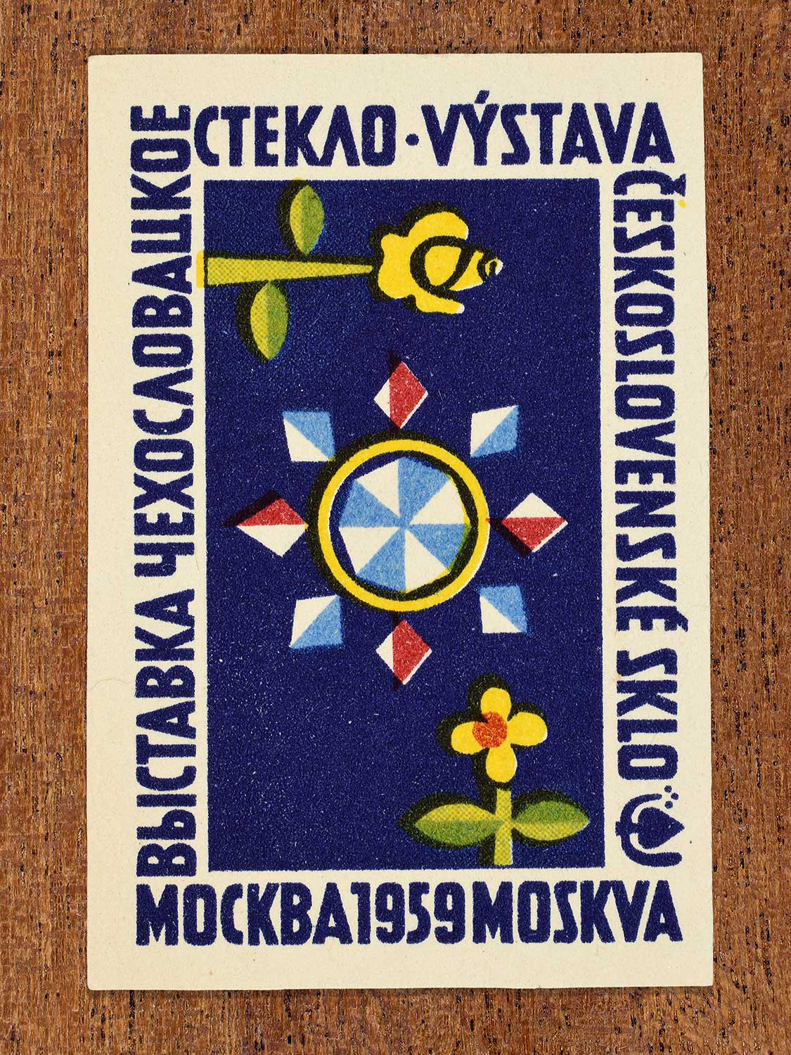 Vintage 1959 Czechoslovak matchbox label showing a central decorative circular starburst in red, white, blue and yellow, framed by delicate yellow flowers on dark blue. Produced by an unknown factory to promote Czechoslovak artistic glass design at an international cultural event.