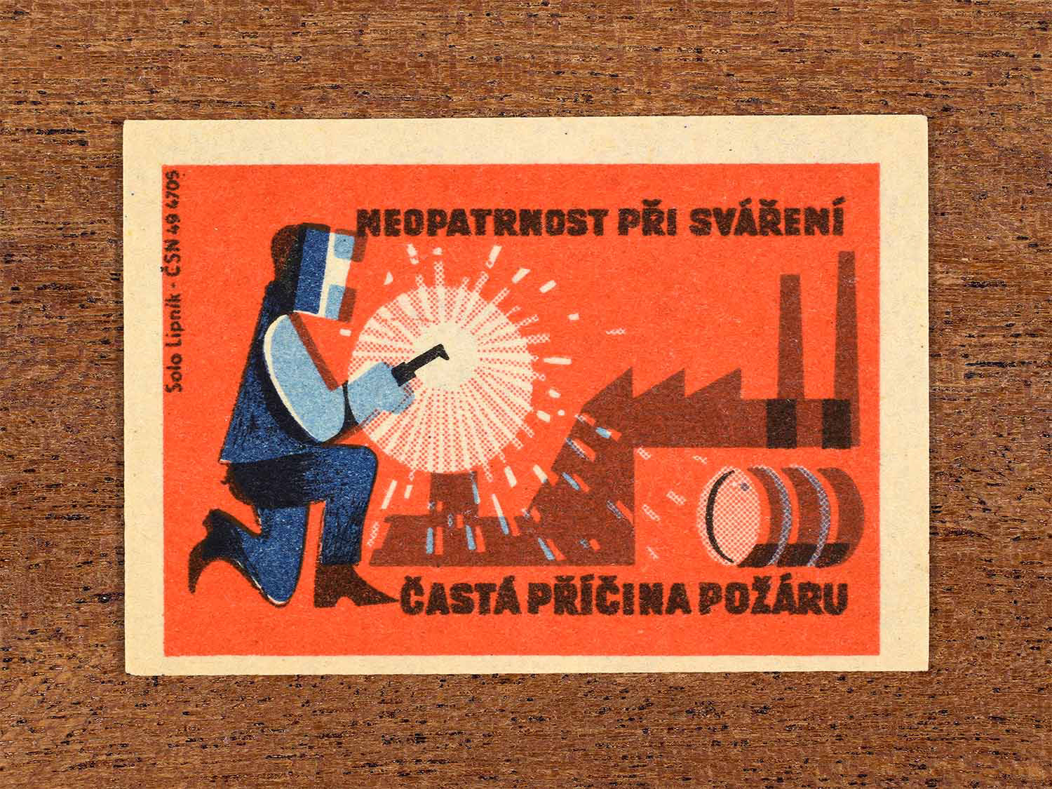 Vintage 1964 Czech matchbox label showing a worker welding in an industrial setting, in blue and red colours. Produced by the Solo Lipník match factory to warn that careless welding is a common cause of fires.