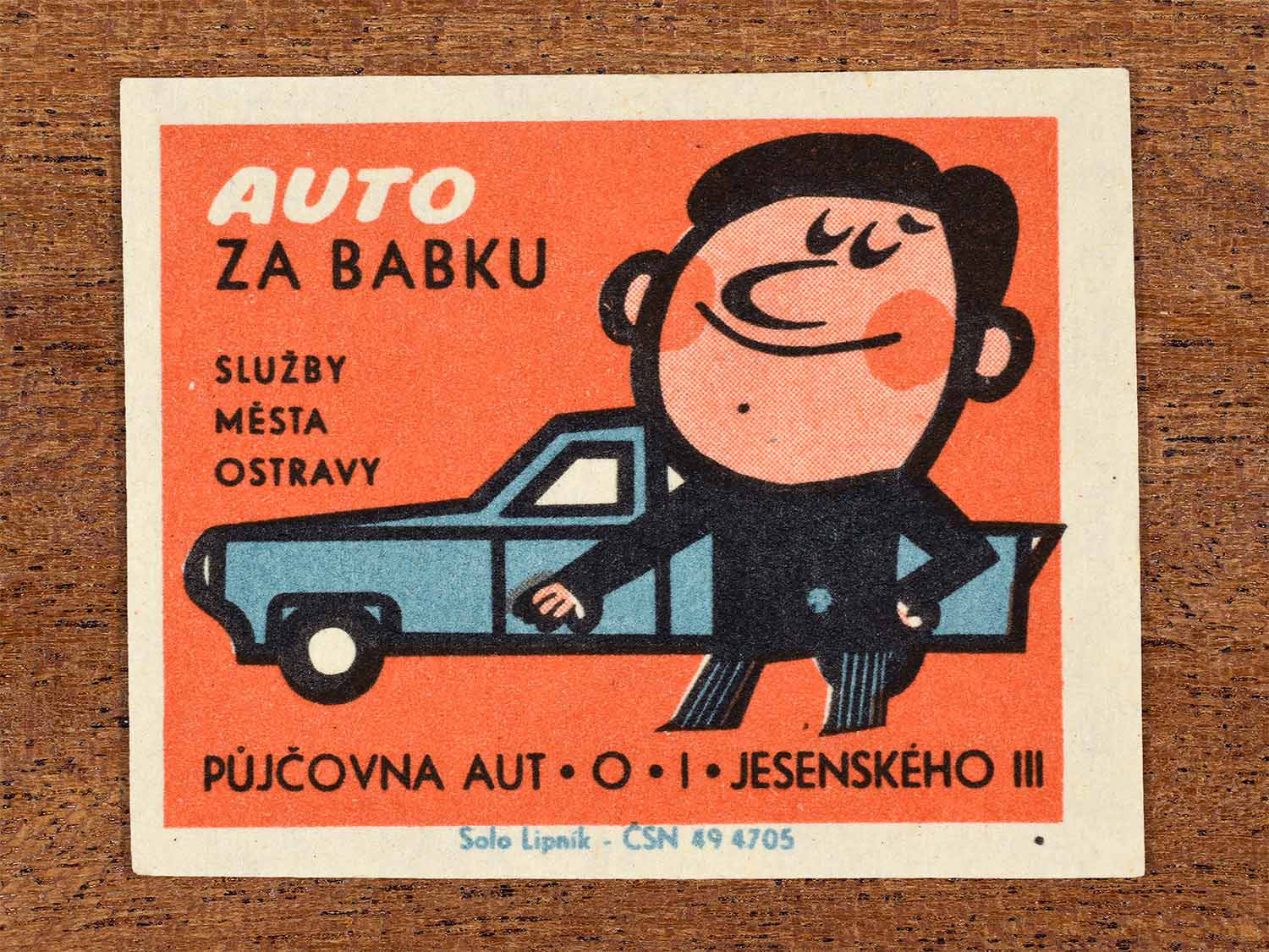 Vintage 1967 Czech matchbox label showing a smiling cartoon man driving a blue car, on a red background. Produced by the Solo Lipník match factory to advertise municipal car rental services.
