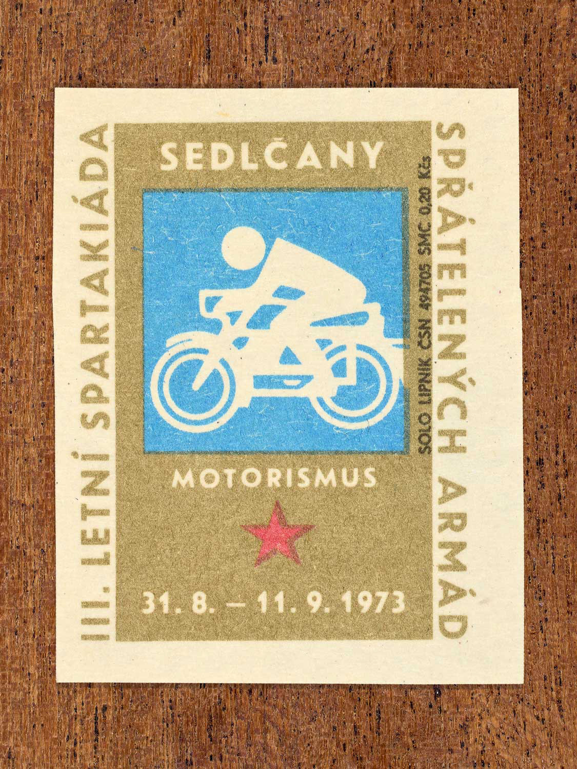 Vintage 1973 Czech matchbox label showing a simplified white motorcycle rider on a blue and gold background. Produced by the Solo Lipník match factory to promote physical training and demonstrate collective military readiness among allied armies.