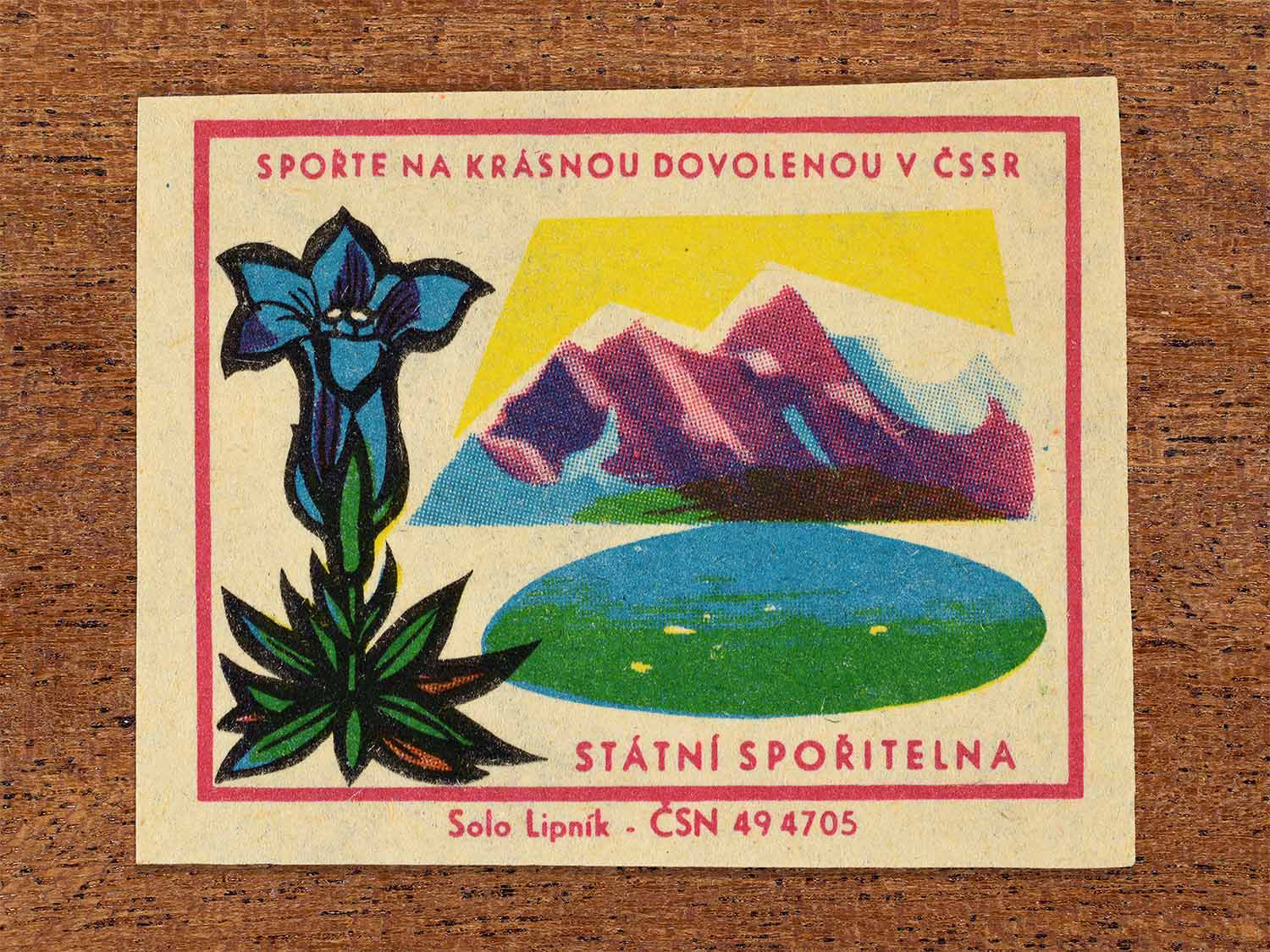 Vintage 1968 Czech matchbox label showing an alpine flower beside a mountain lake and purple mountain range. Produced by the Solo Lipník match factory to promote saving money for holidays in the Czechoslovak Socialist Republic.
