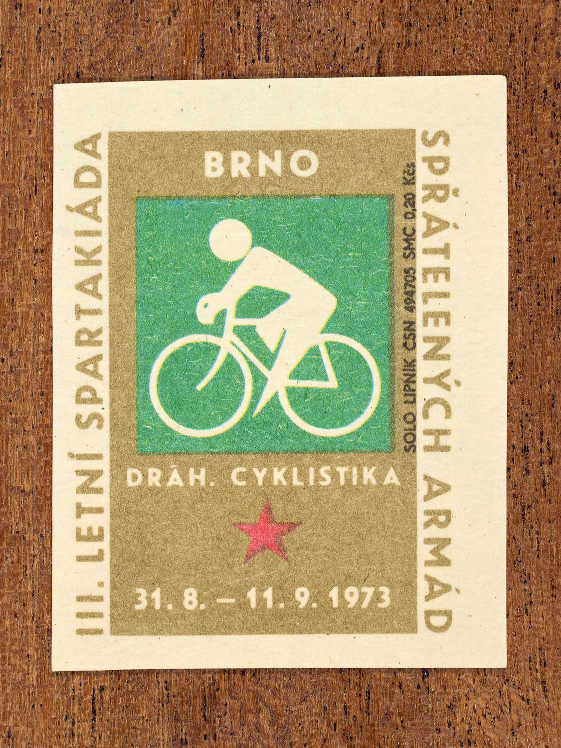 Vintage 1973 Czech matchbox label showing a simplified white cyclist on a green and gold background. Produced by the Solo Lipník match factory to promote physical training and demonstrate collective military readiness among allied armies.