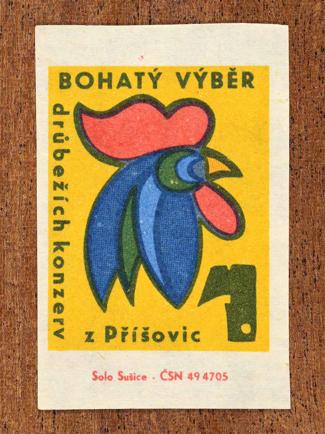 Vintage c. 1966 Czech matchbox label showing a stylised rooster head advertising a wide selection of poultry preserves from Příšovice in bold yellow, red and blue. Produced by the Solo Lipník match factory to promote variety in preserved foods.