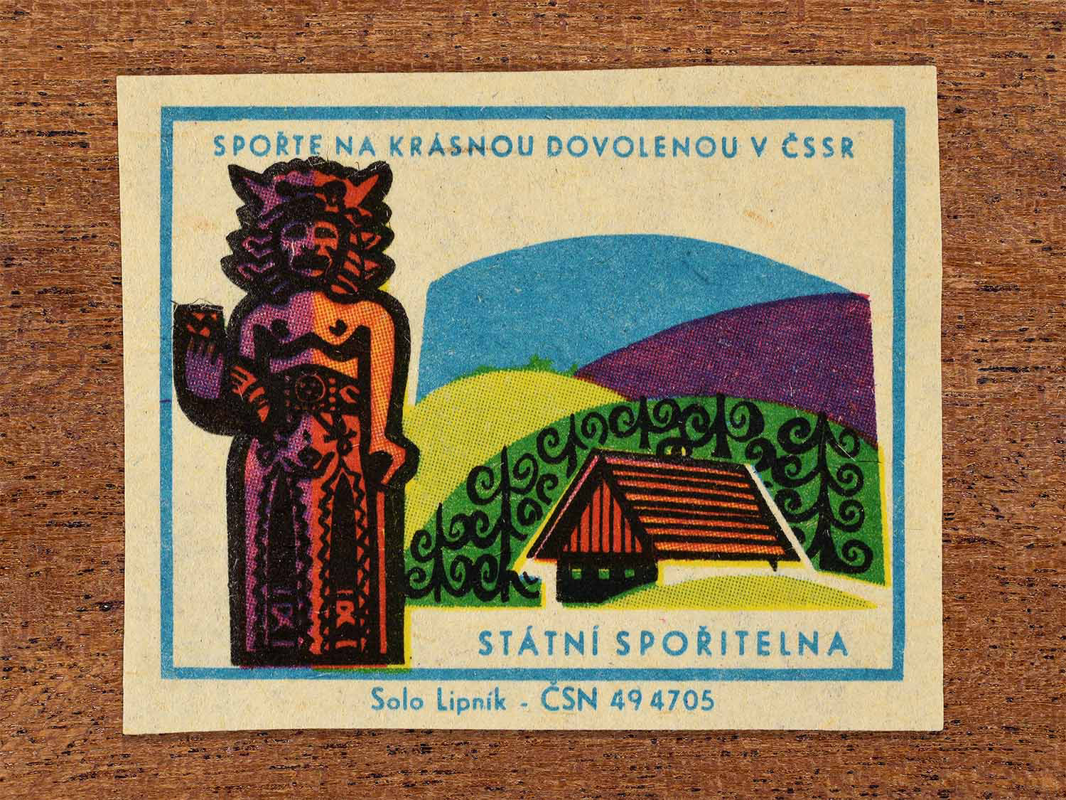 Vintage 1968 Czech matchbox label showing a carved folk statue beside a rural cottage and rolling hills. Produced by the Solo Lipník match factory to promote saving money for holidays in the Czechoslovak Socialist Republic.