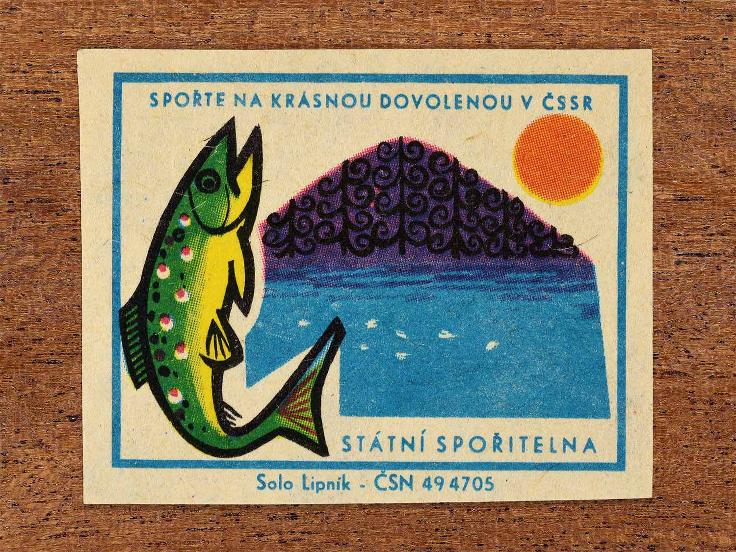Vintage 1968 Czech matchbox label showing a large stylised fish beside a lake and wooded hill at sunset. Produced by the Solo Lipník match factory to promote saving money for holidays in the Czechoslovak Socialist Republic.