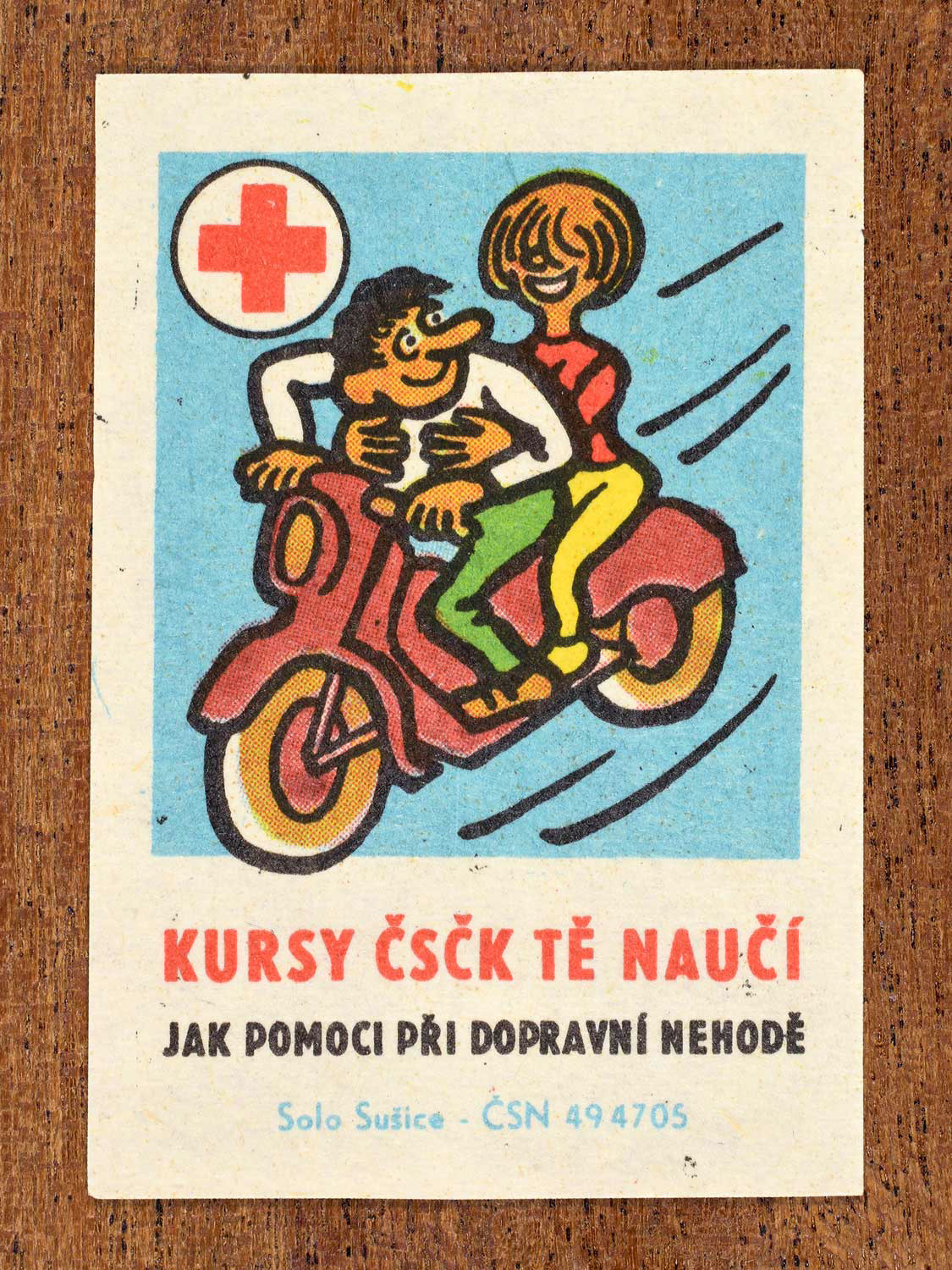 Vintage c. 1966 Czech matchbox label showing a man and woman riding a red motorbike. Produced by the Solo Sušice match factory to promote first-aid education and traffic emergency assistance in cooperation with the Czechoslovak Red Cross.