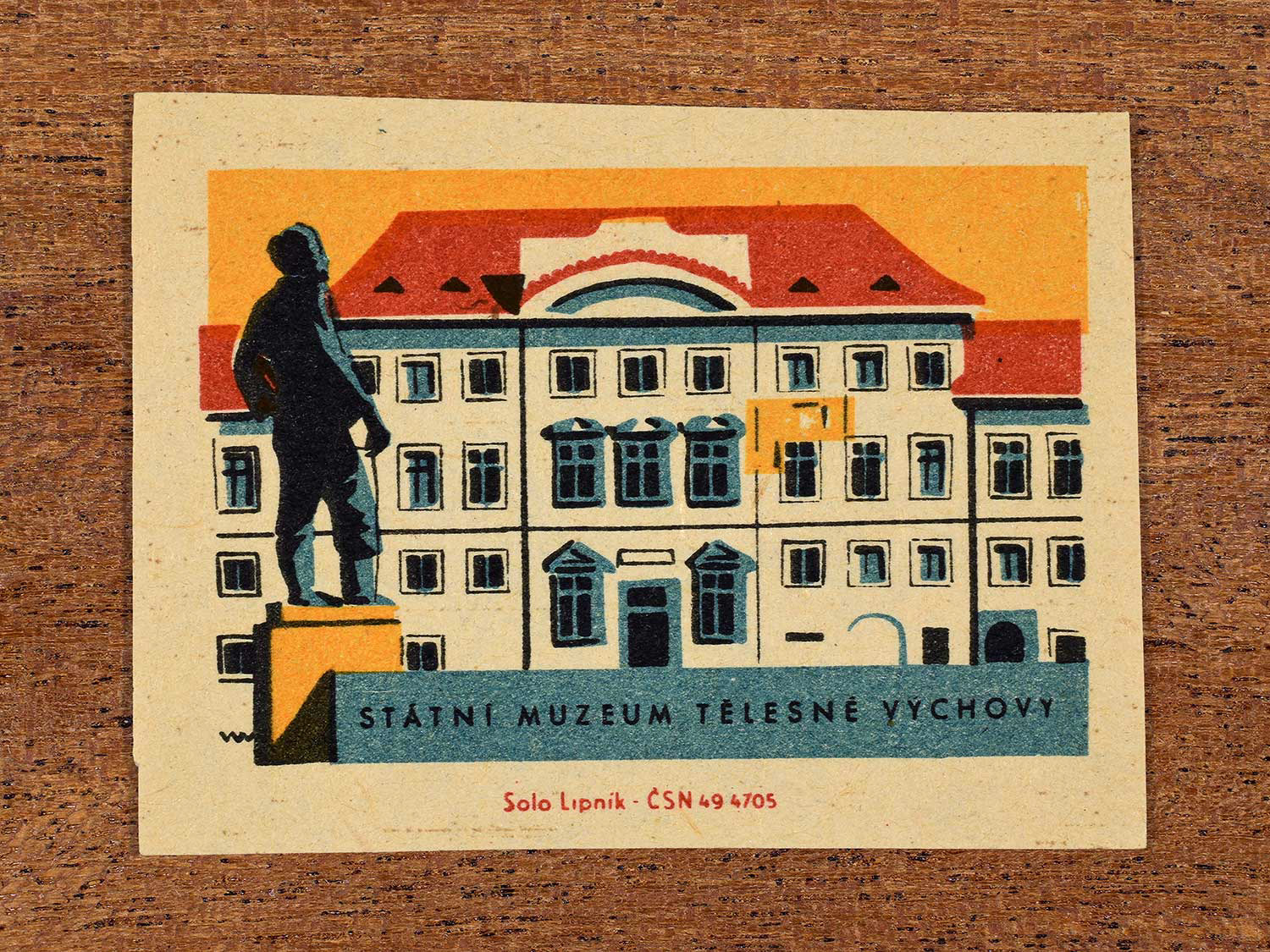 Vintage 1964 Czechoslovakia matchbox label showing the Státní Muzeum Tělesné Výchovy (State Museum of Physical Education) in yellow, red and teal colours. Produced by the Solo Lipník match factory to promote domestic historical heritage and municipal institutions across the state.mu