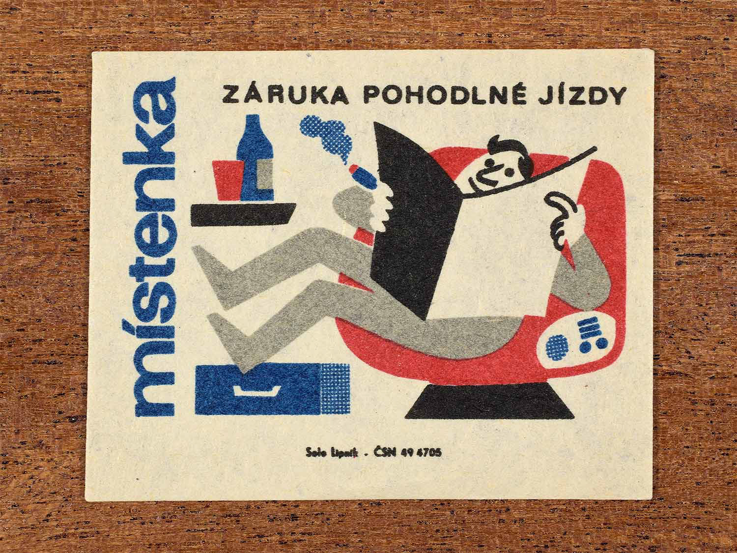 Vintage c. 1964 Czech matchbox label showing a relaxed passenger reclining comfortably with refreshments, illustrated in blue, red and black. Produced by the Solo Lipník match factory to promote seat reservations for comfortable train travel.