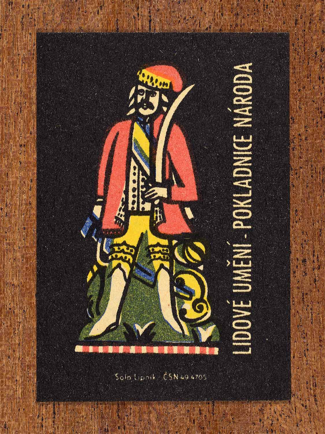 Vintage c. 1962 Czech matchbox label showing a stylised male folk figure in traditional dress holding a ceremonial sword, rendered in bold folkloric colours. Produced by the Solo Lipník match factory to celebrate folk art as a national cultural heritage.