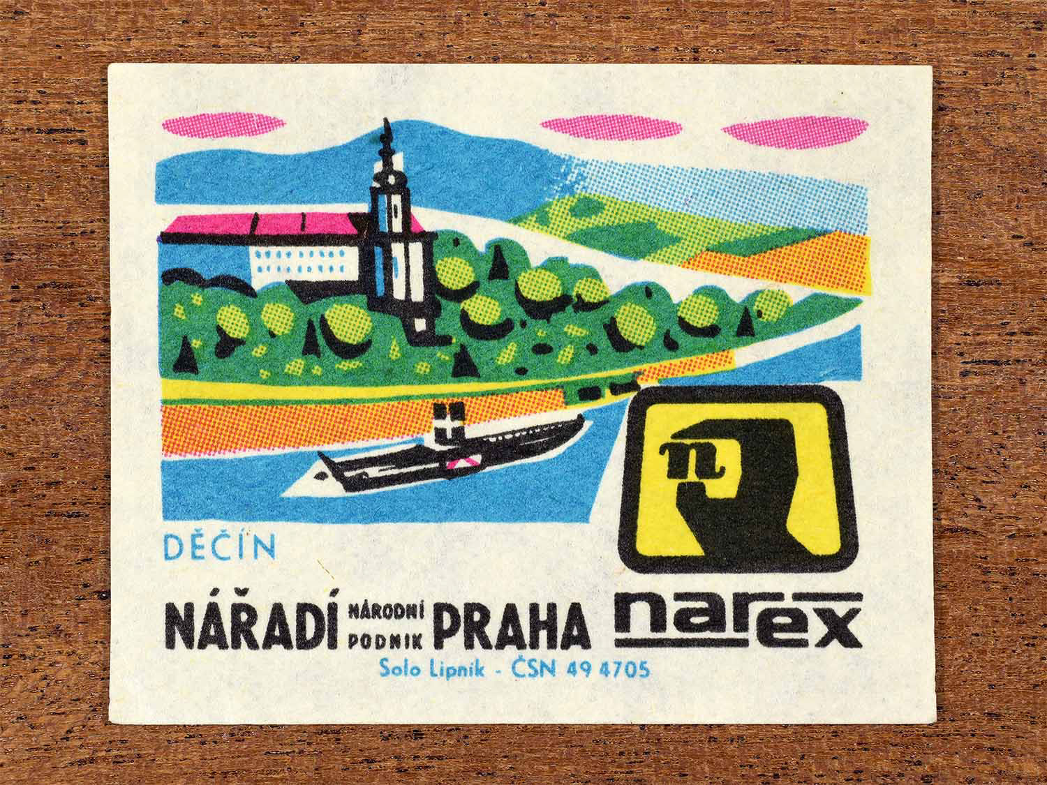 Vintage 1967 Czech matchbox label showing a riverside town with castle architecture and a boat on the river in multiple bright colours. Produced by the Solo Lipník match factory to promote Děčín and Narex, the national tool enterprise in Czechoslovakia.